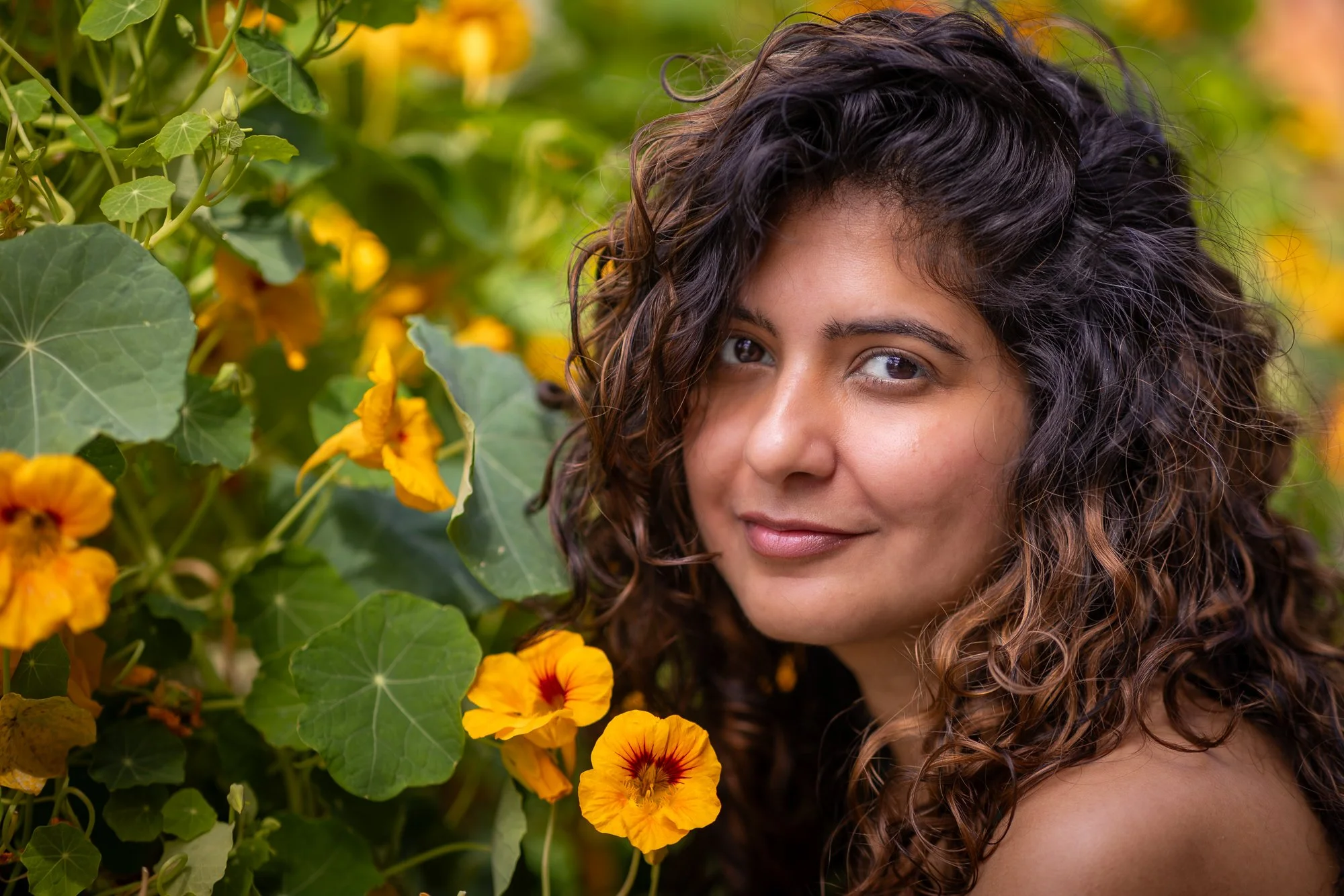 A woman with curly dark brown hair and warm eyes surrounded by yellow and orange flowers and green leaves.