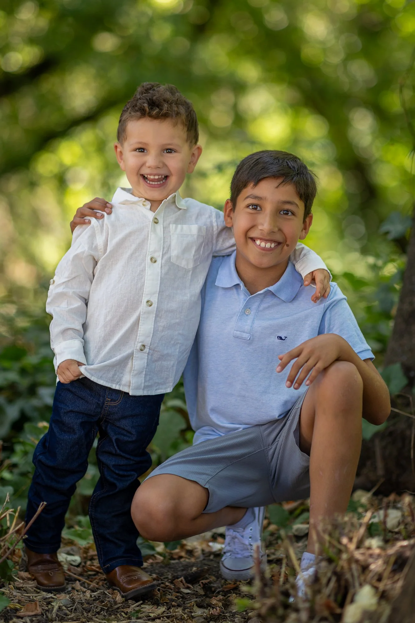 Two young boys outdoors among trees; one is standing and the other is crouching, with their arms around each other's shoulders, smiling happily.