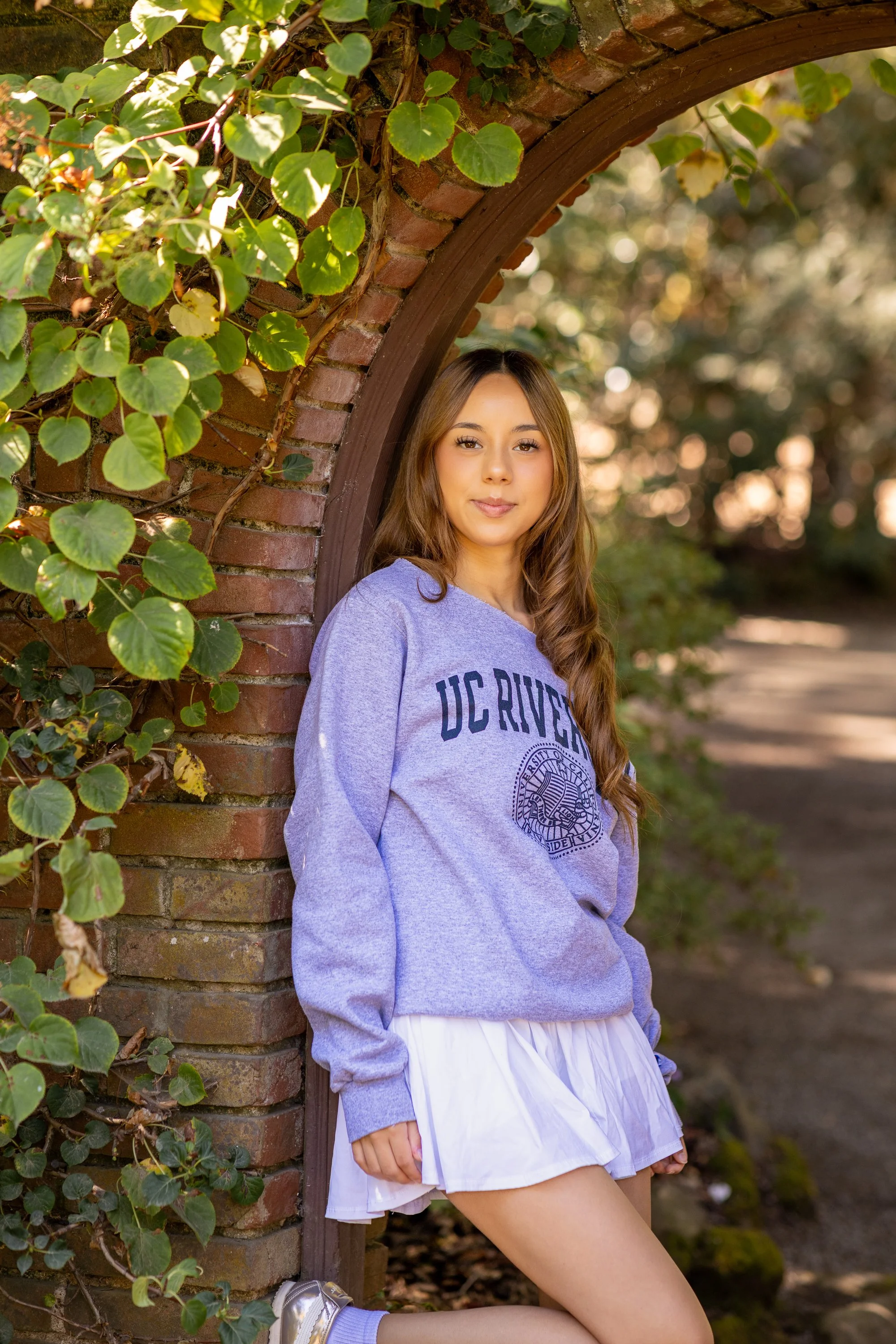 A young woman in a purple hoodie and white skirt leaning against a brick archway covered with green leaves during sunset.