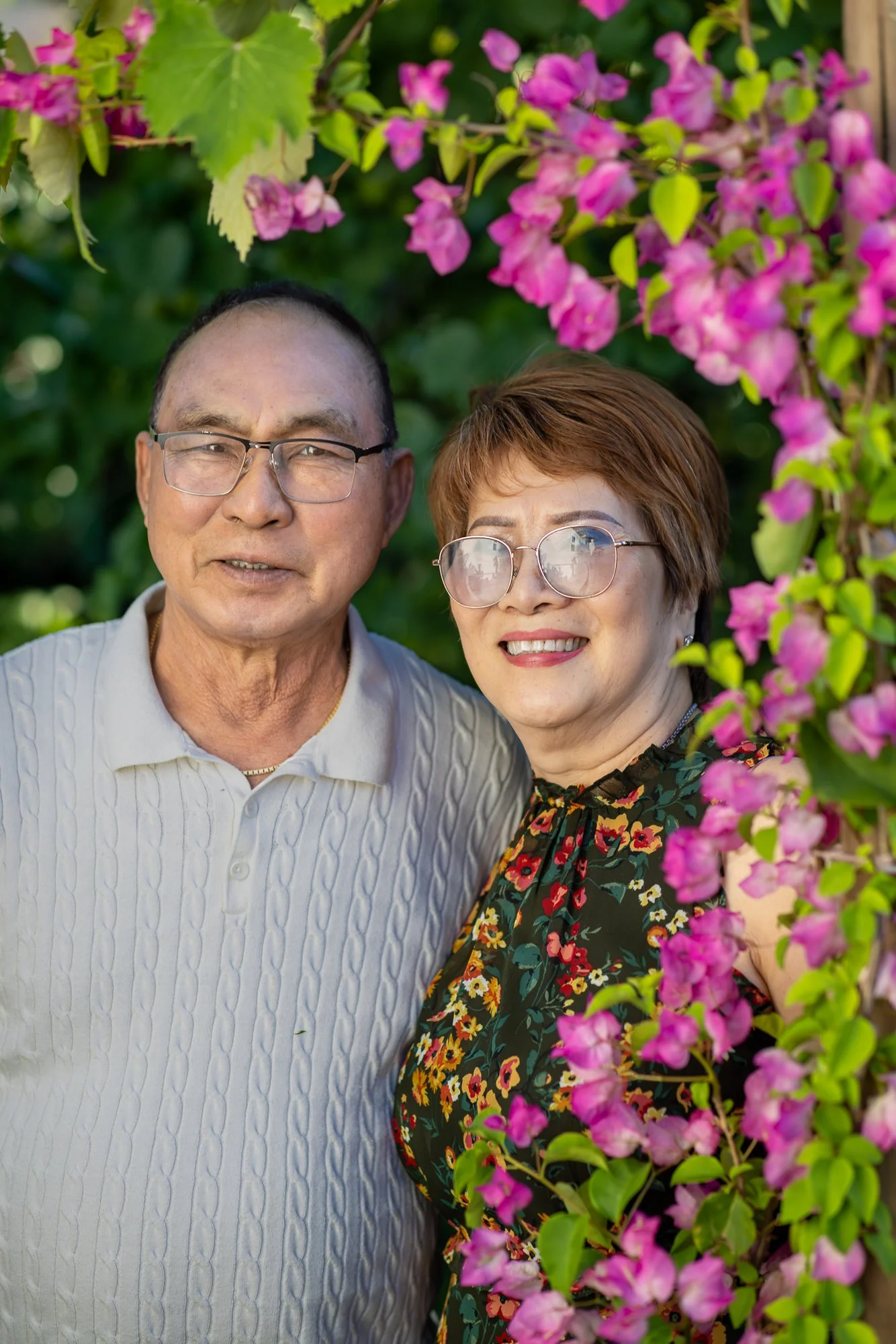 A smiling middle-aged couple standing outdoors, surrounded by green leaves and pink flowers.