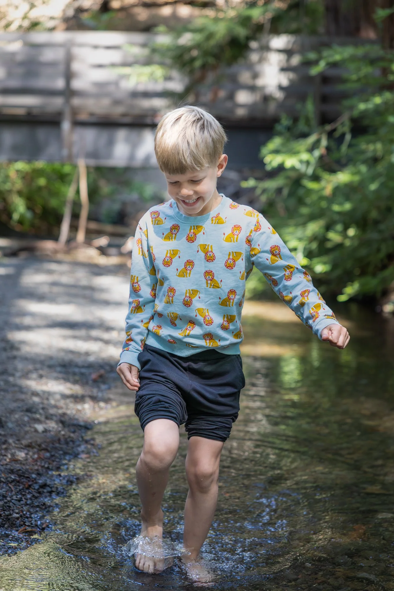A young boy with blonde hair smiling and walking through a shallow stream in a wooded outdoor area, wearing a long-sleeved shirt with lion print and dark shorts.