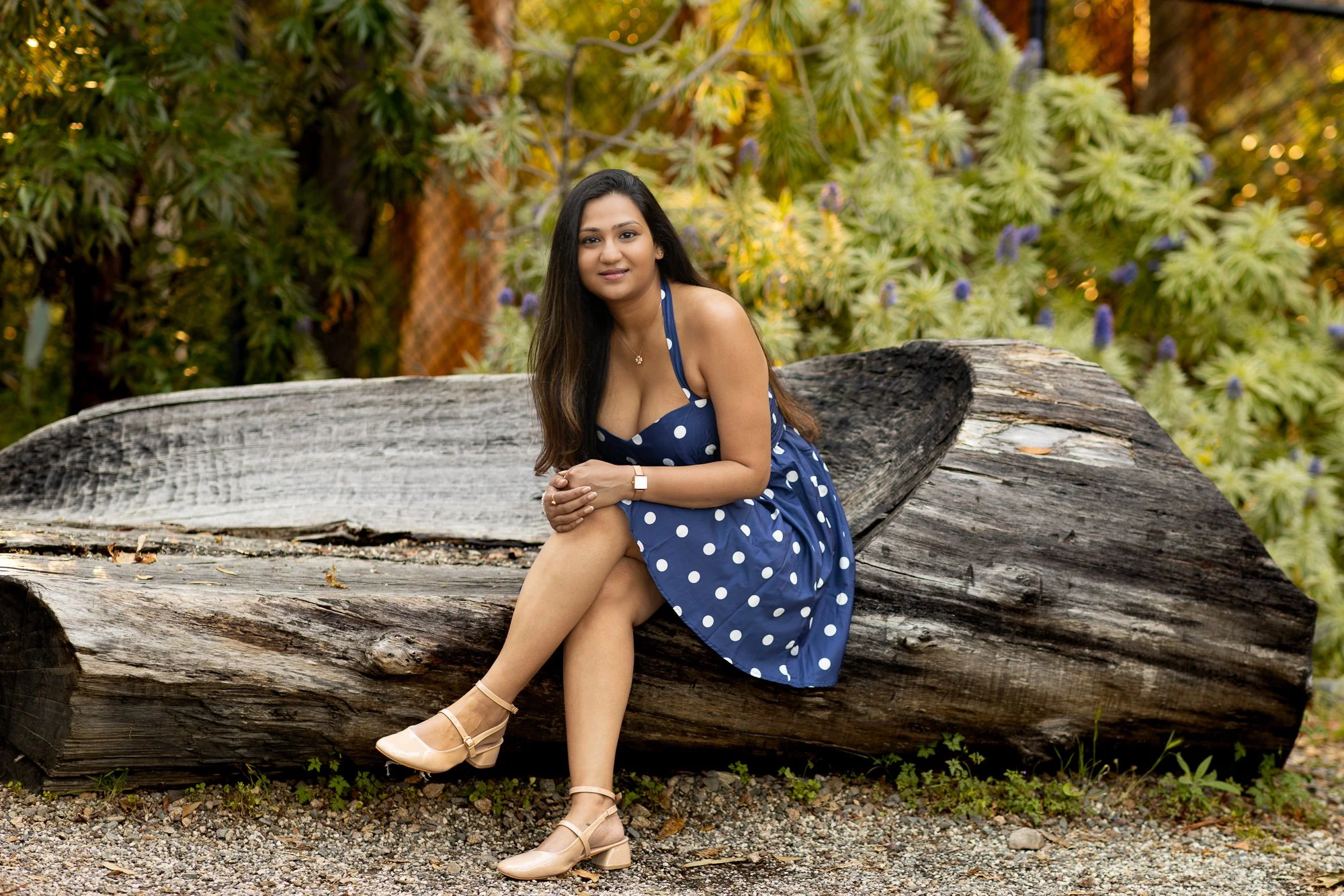 A woman wearing a blue polka dot dress and beige heels sitting on a large fallen log in a park with green and yellow foliage in the background.