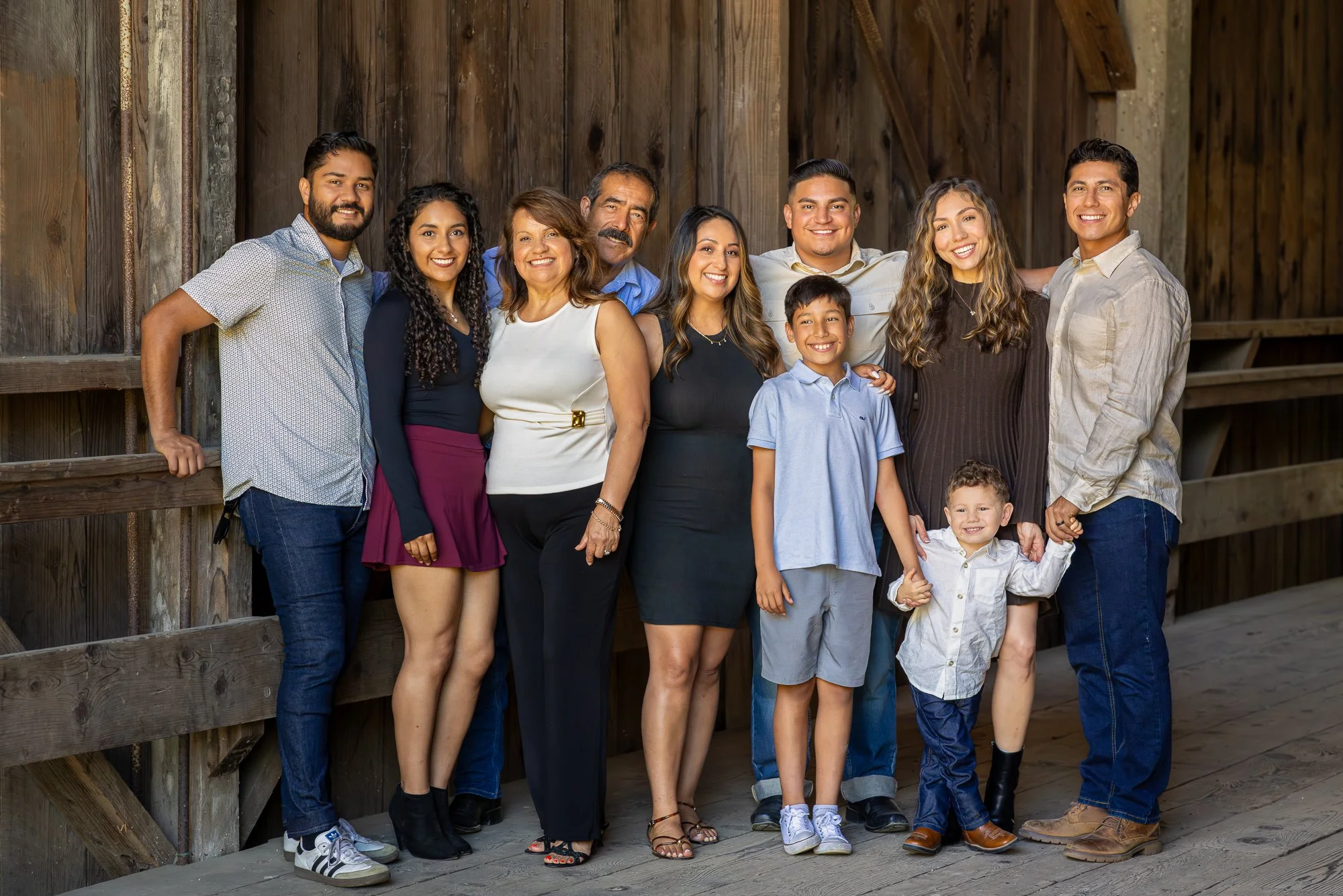 A diverse group of people, including adults and children, standing together outdoors against a rustic wooden wall, smiling and looking at the camera.