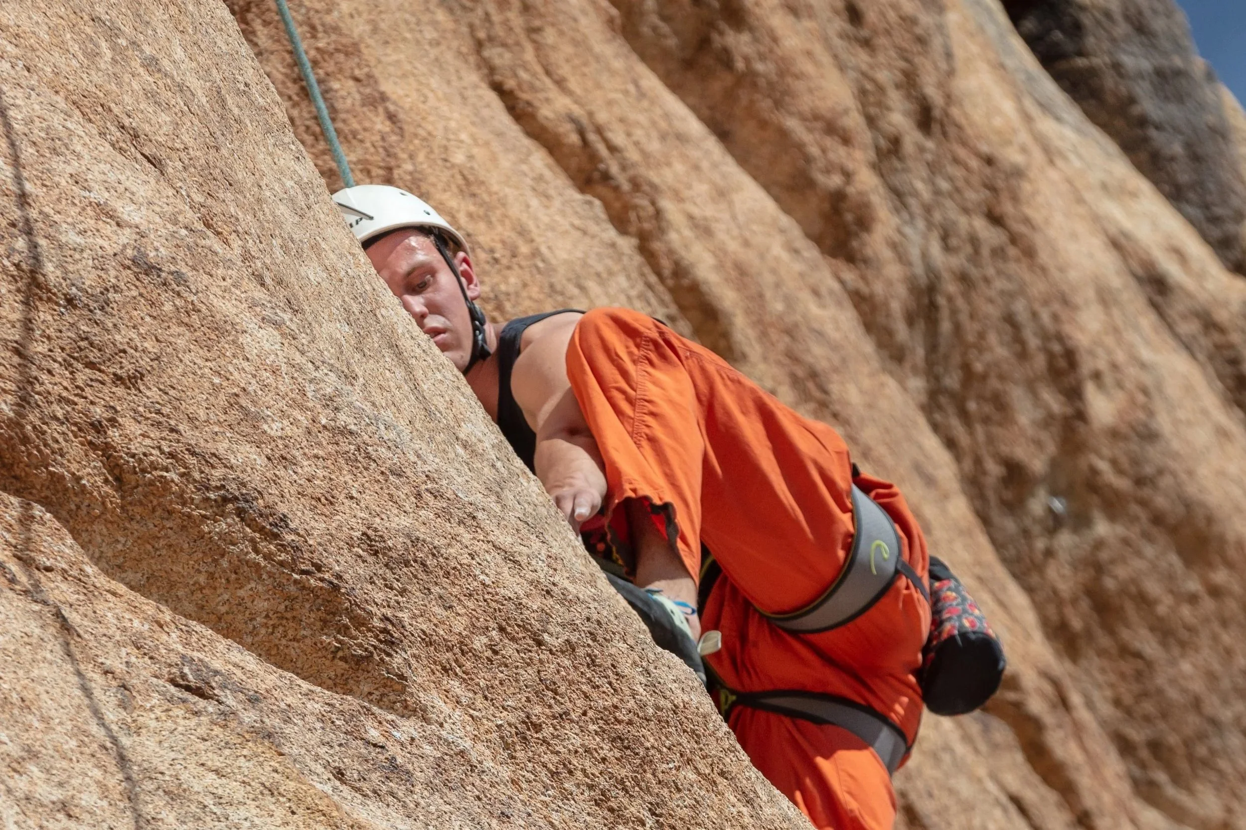 A man wearing a white helmet and orange climbing pants is rock climbing on a steep, textured stone wall.