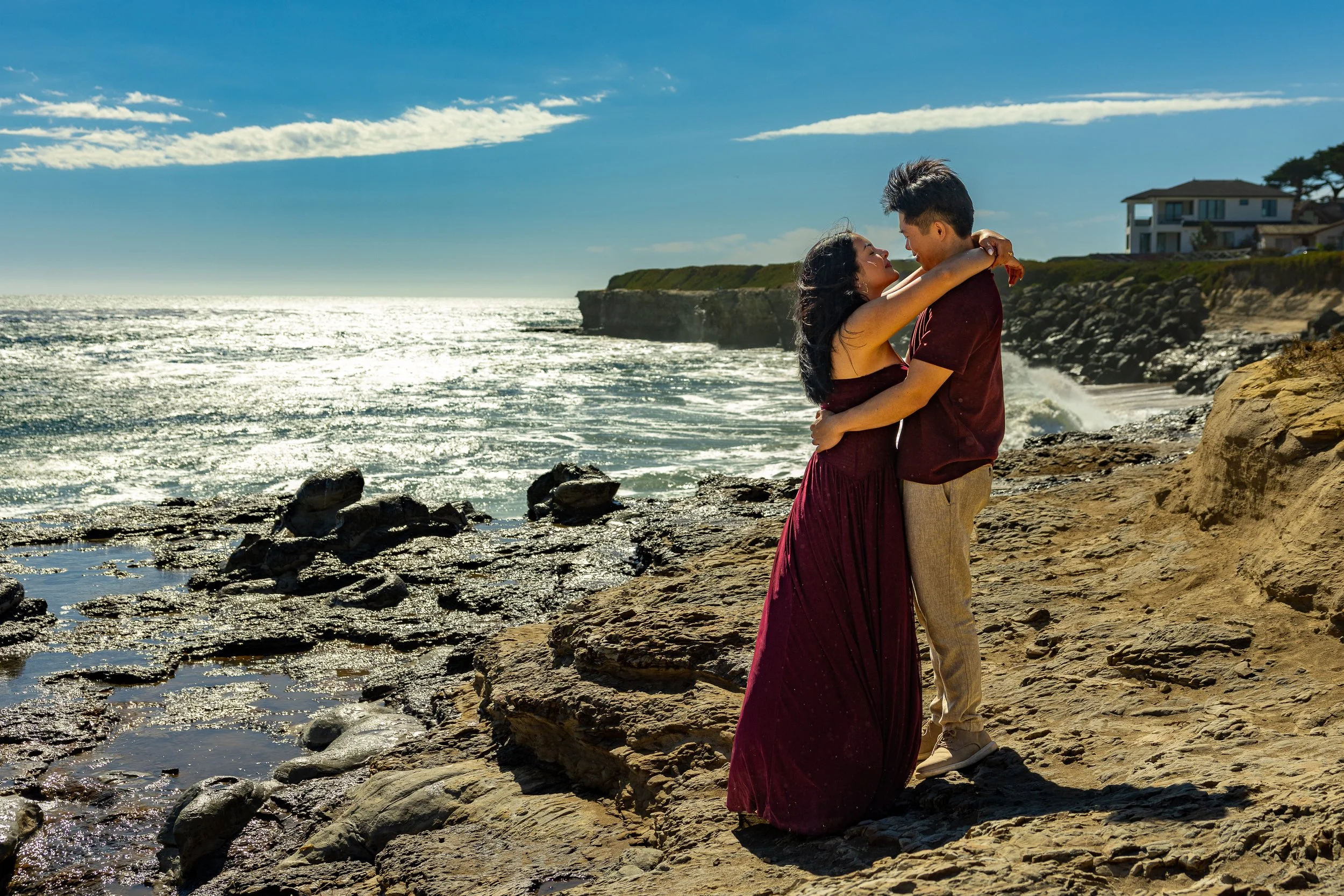 A couple embracing on a rocky beach with the ocean and houses on a cliff in the background during sunny weather.