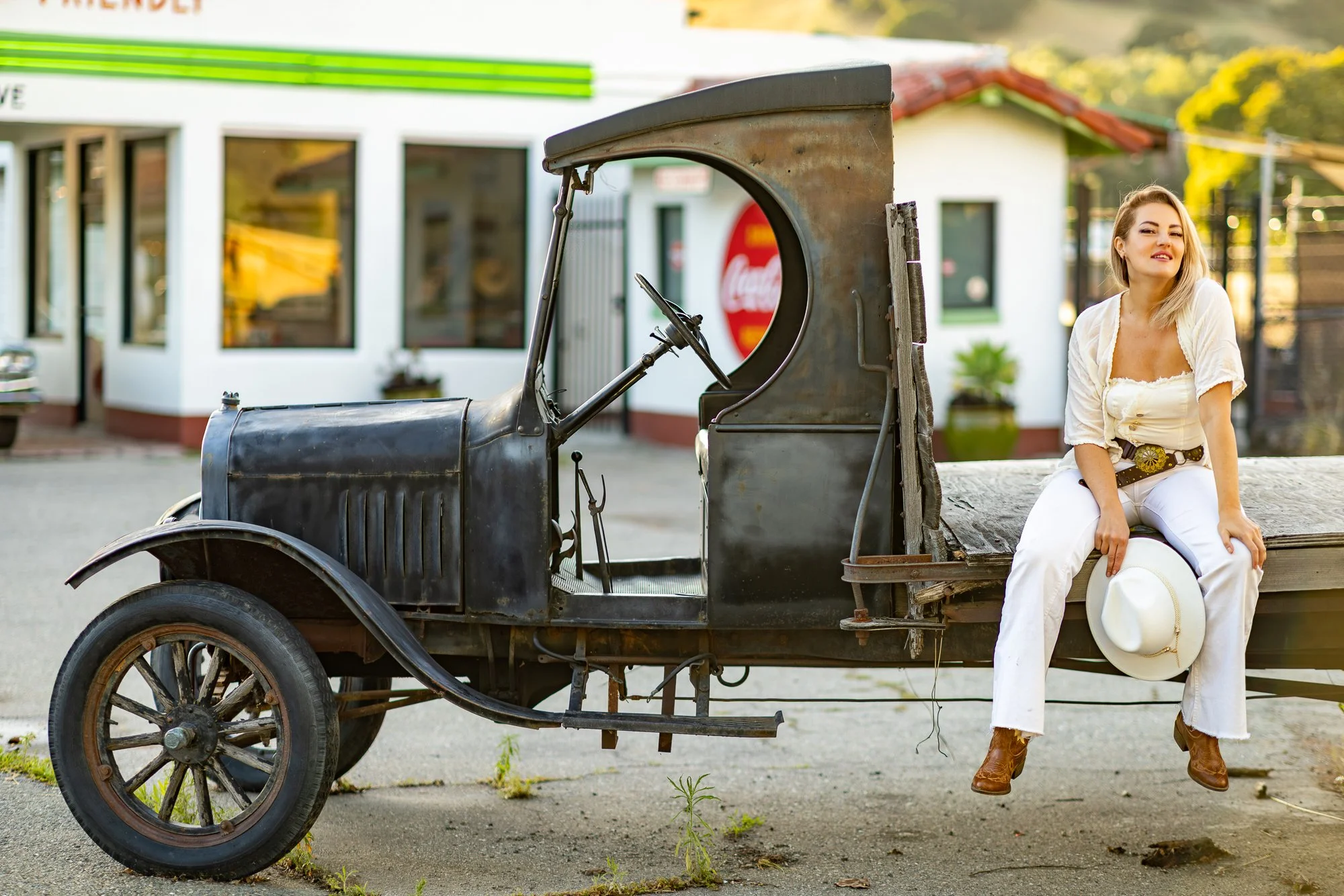 A woman in white outfit sitting on the back of an old, rusty pickup truck with a wooden bed, holding a white hat and a straw hat, with a small house and trees in the background.