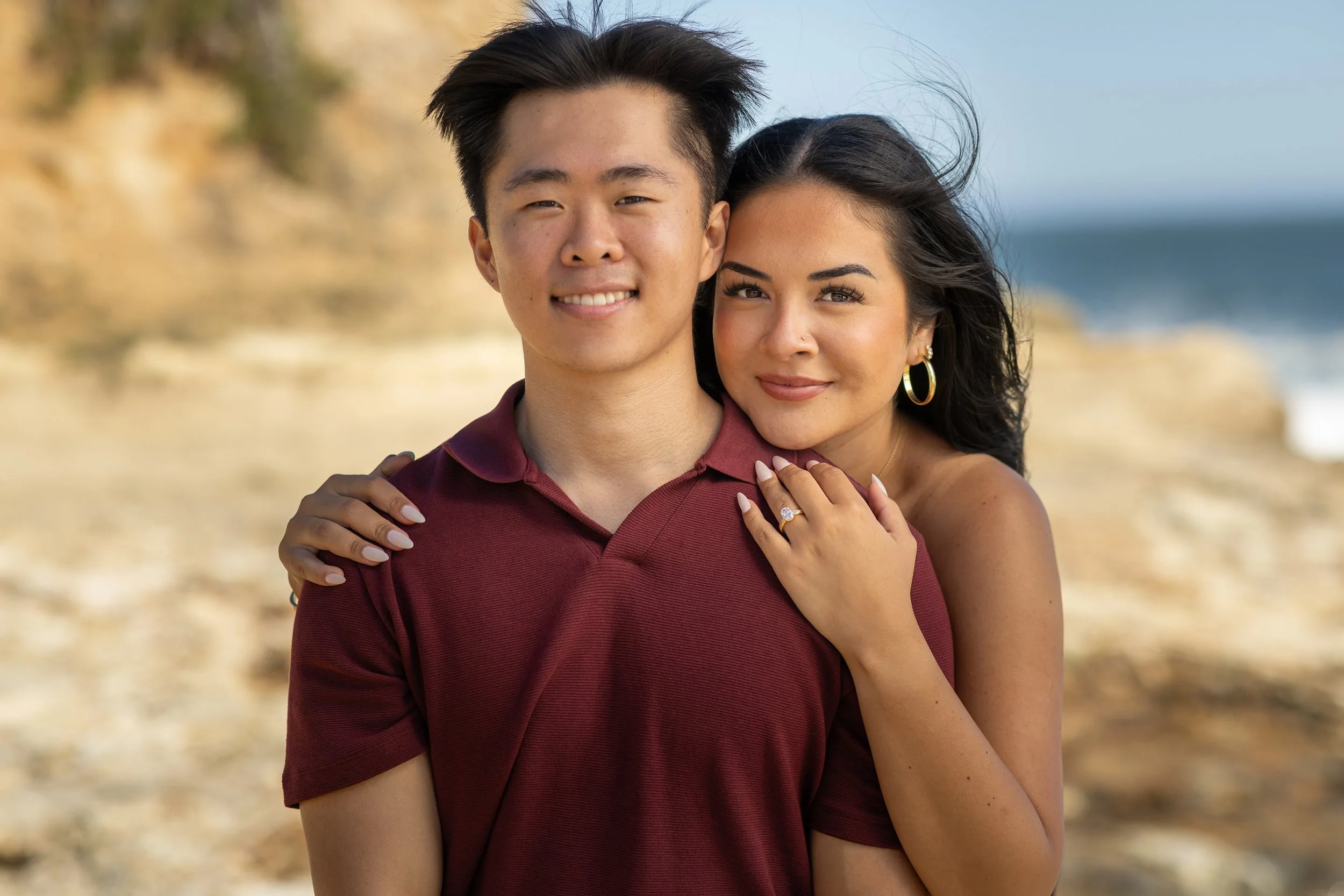A young man and woman are standing close together on a beach, smiling at the camera. The woman is embracing the man with her hands on his shoulder, showing rings on her fingers. The background features a sandy beach with cliffs and ocean waves under 
