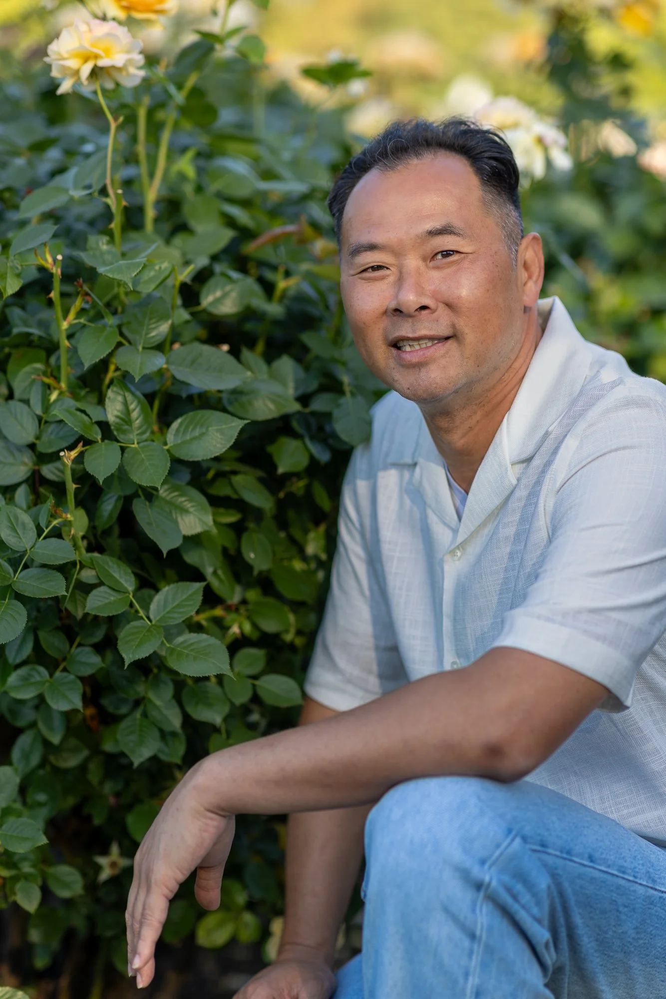 A man with short dark hair and a friendly smile, sitting outdoors among green bushes and white flowers, wearing a white shirt and light blue jeans.