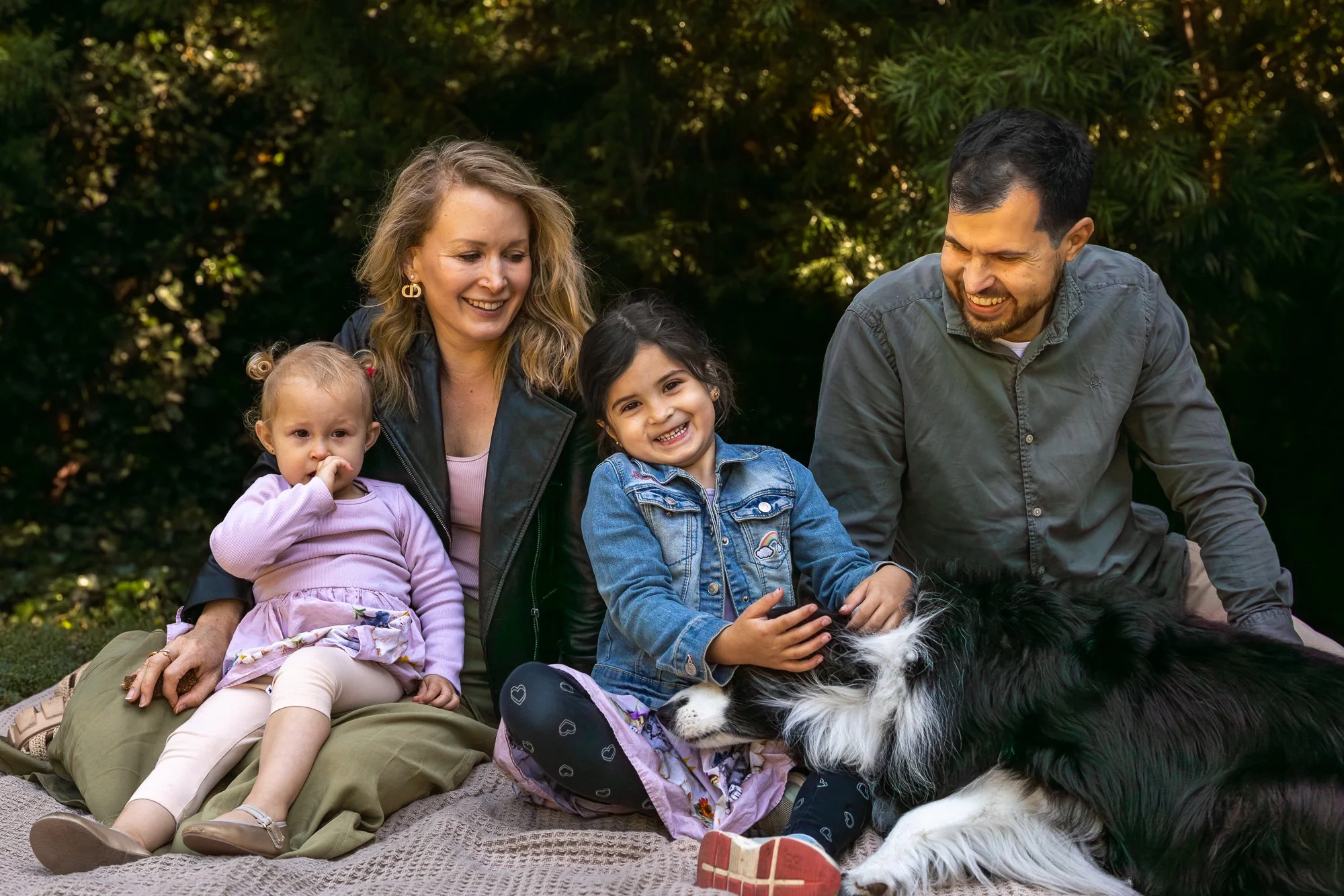 A family of four with two young girls and a man and woman sitting outdoors on a blanket among green foliage, smiling and playing with a black and white border collie dog.