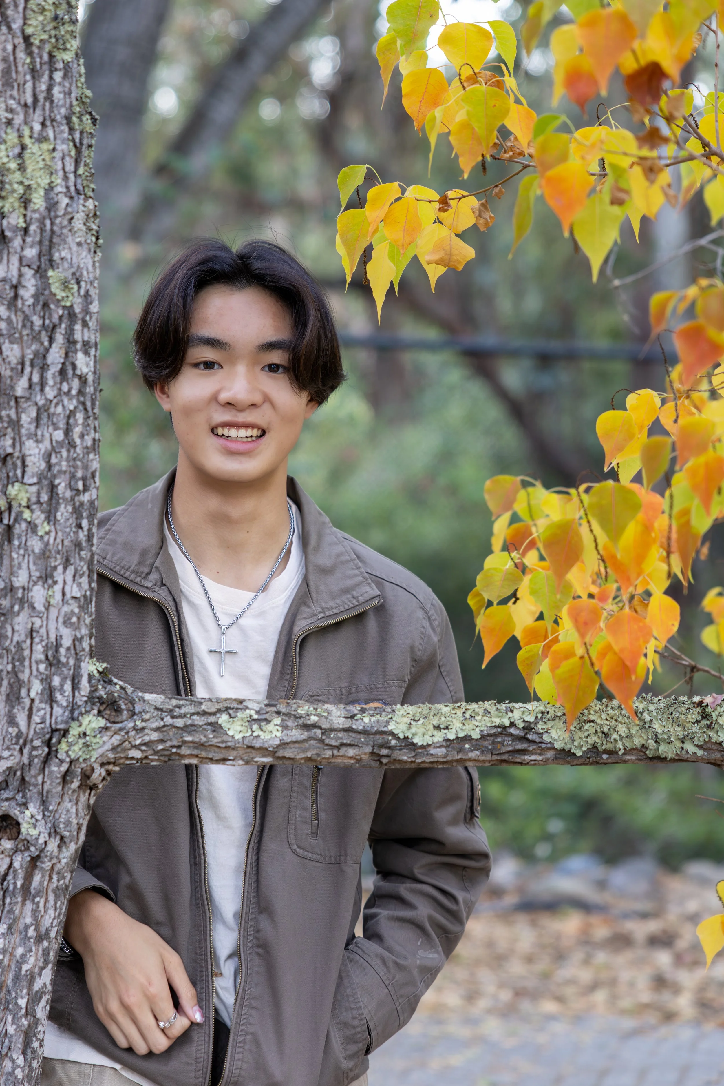 A young man standing outdoors near a tree with autumn-colored leaves, smiling and posing with one hand in his pocket.