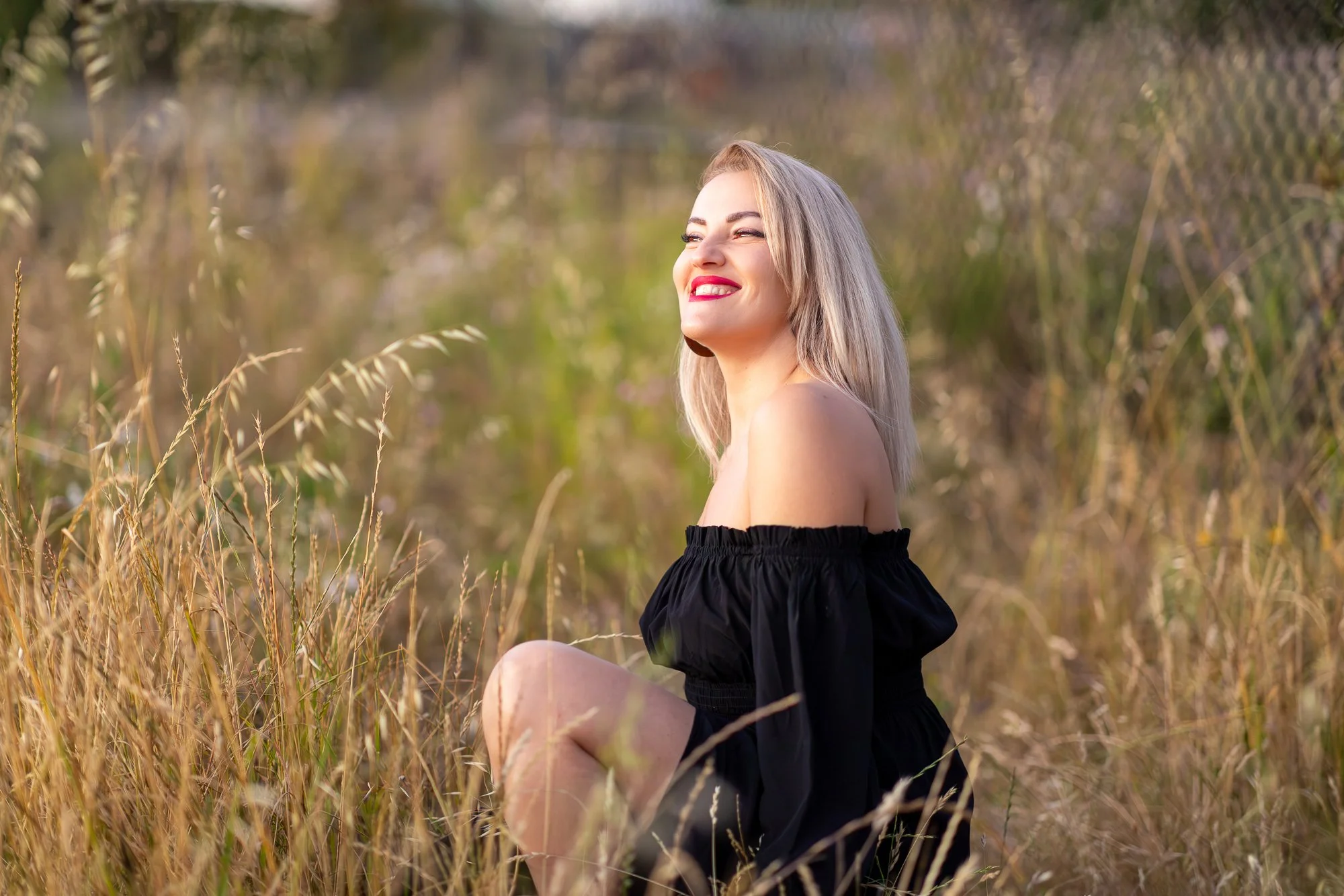 A woman sitting in a field of tall, golden grasses, wearing a black off-the-shoulder dress, smiling and looking to the side.