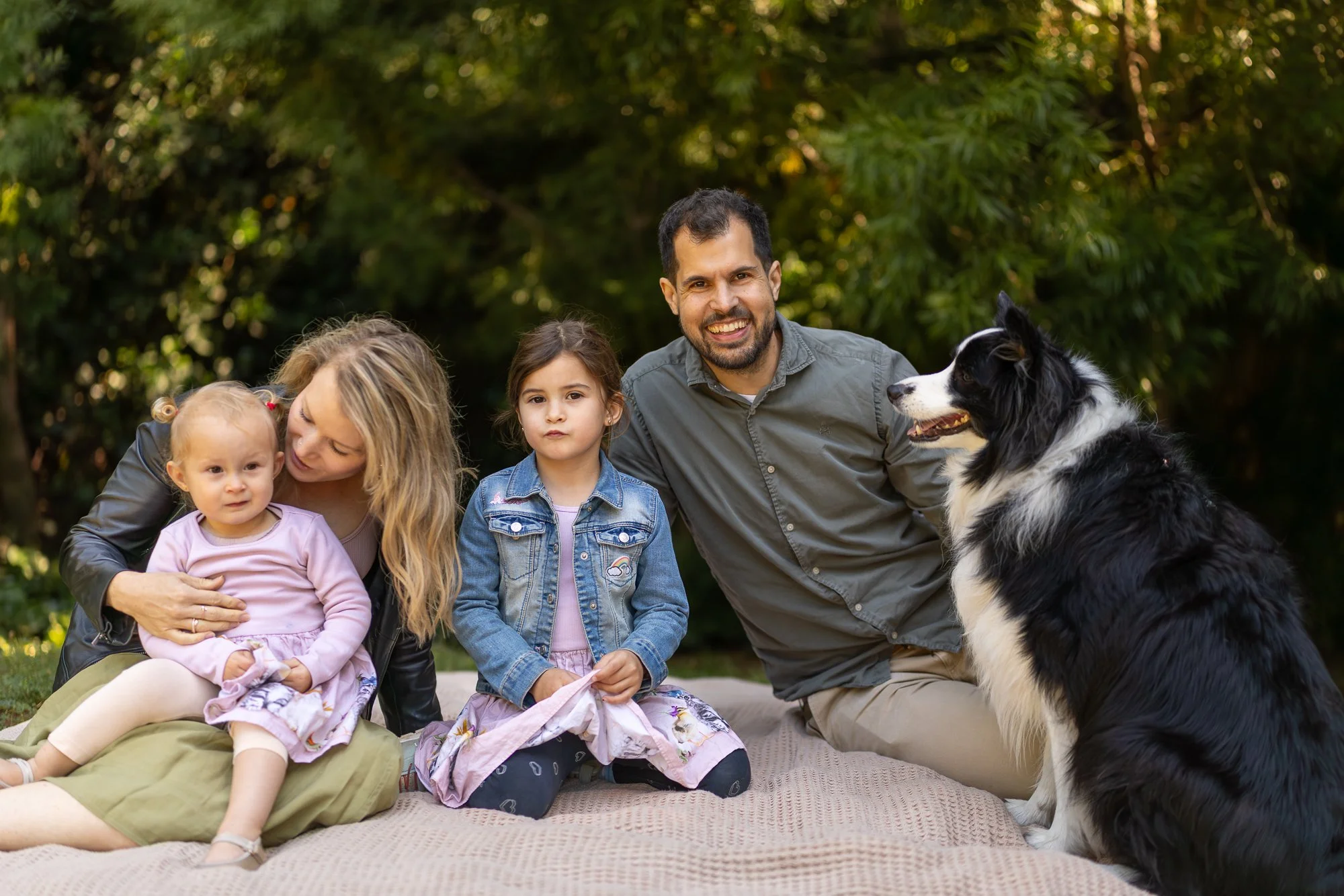 A family of four with a black and white dog outdoors on a sunny day. The family includes a woman, a man, and two young girls. They are sitting on a blanket on the grass with trees in the background.