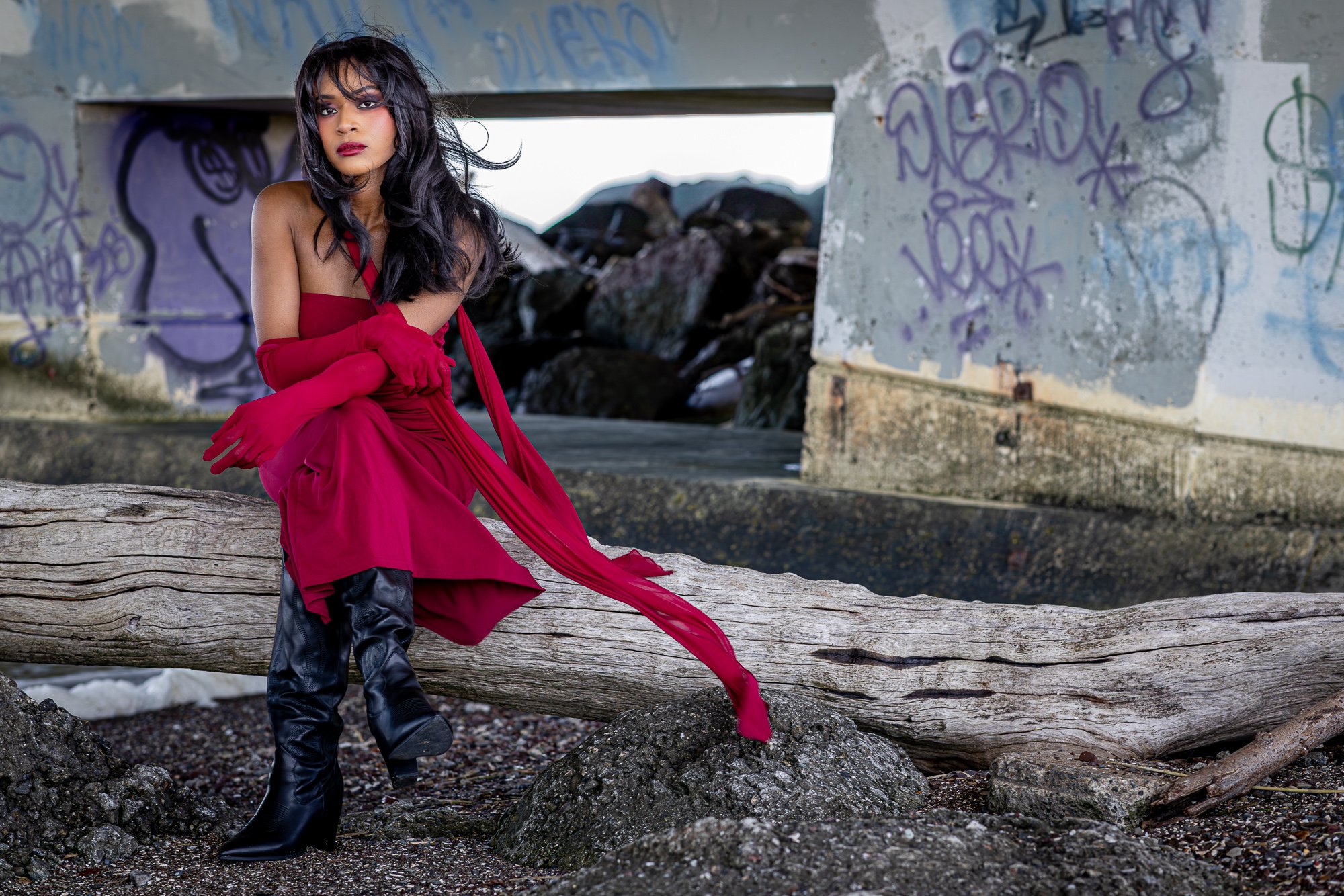 Woman in crimson gown on driftwood log at Dumbarton Shoreline, graffiti bridge abutments, editorial portrait Bay Area
