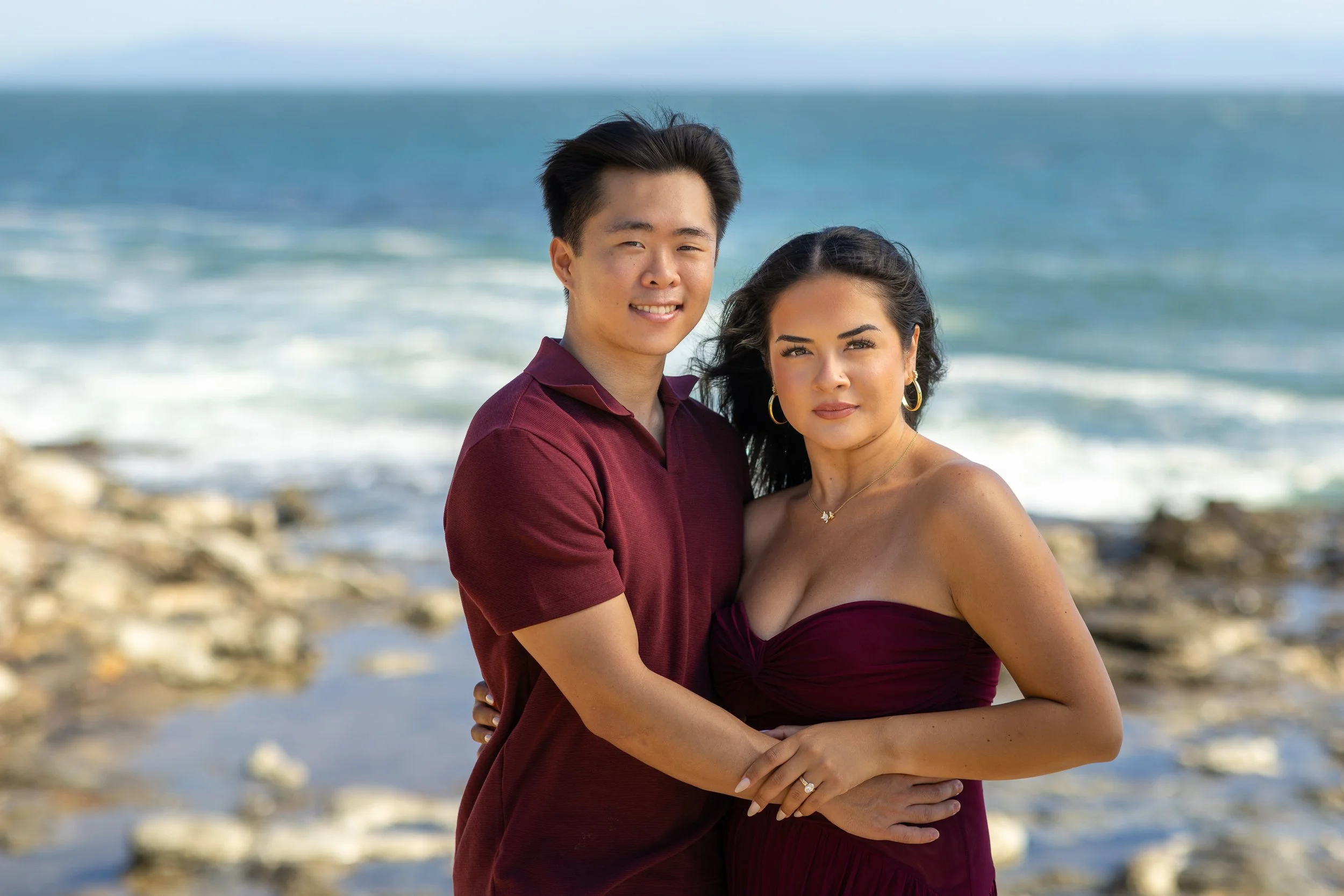 A couple standing close together on a rocky beach near the ocean, smiling at the camera. The man has short dark hair and is wearing a maroon polo shirt, while the woman has long dark hair and is wearing a strapless maroon dress with jewelry.