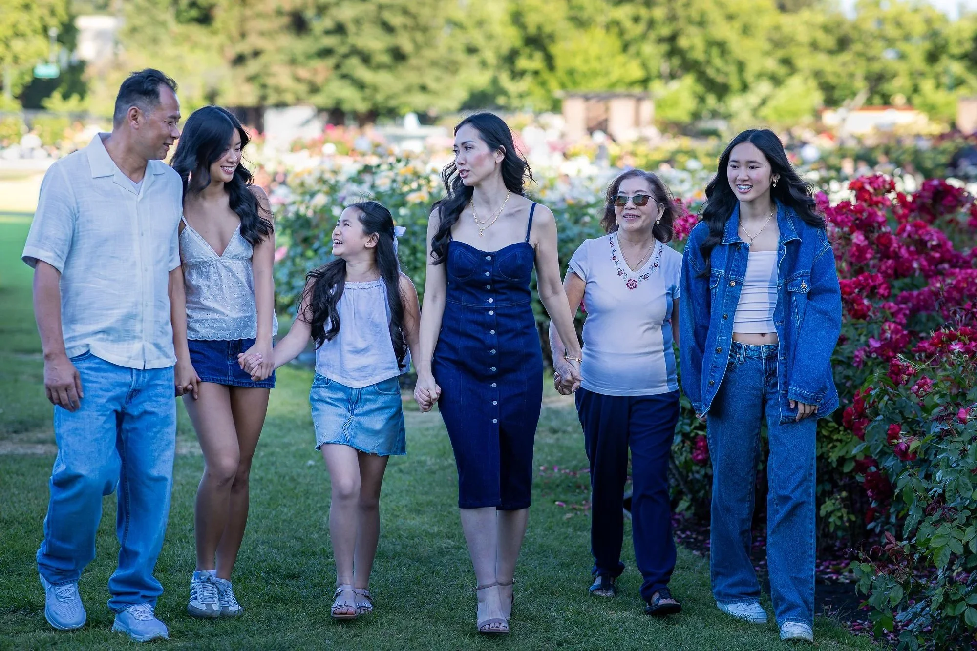 A multi-generational family of six walking together in a park surrounded by colorful flowers and trees on a sunny day.