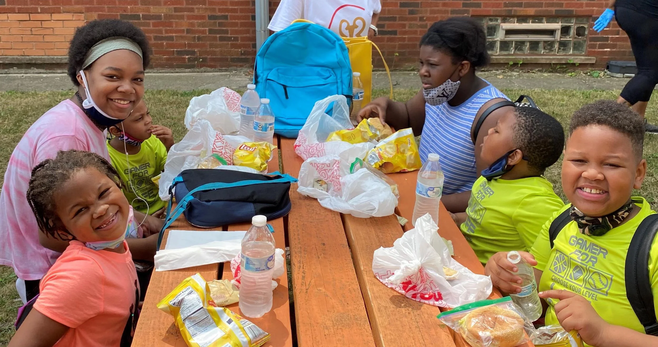 Children and a woman sitting at a picnic table outdoors, eating snacks and drinking water, with backpacks and snack bags on the table.