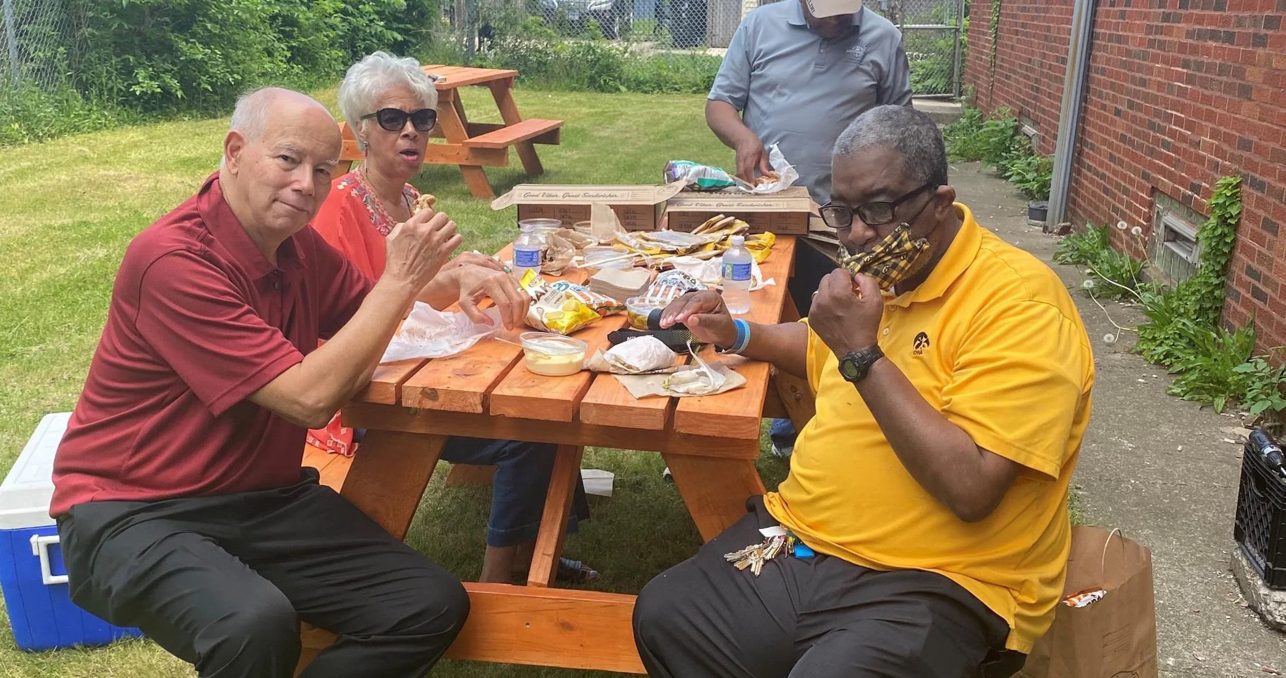 Four people are sitting and standing around a picnic table outdoors, eating and unpacking food. Two men are sitting at the table, one in a maroon shirt and the other in a yellow shirt with a face mask pulled down. A woman and a man are standing at th