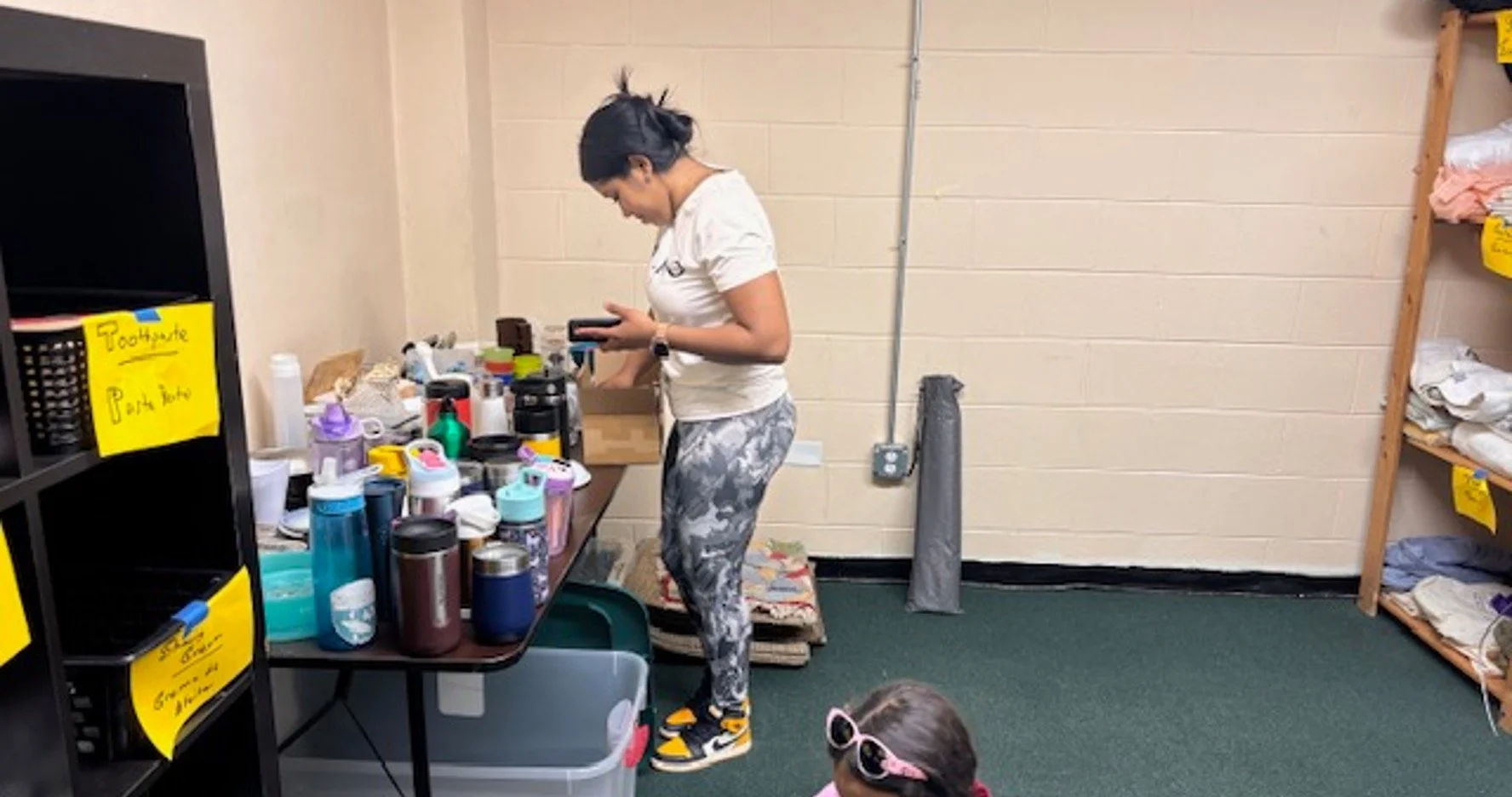 A woman with dark hair tied back, wearing a white t-shirt, patterned leggings, and black and yellow sneakers, looking at her phone while standing at a cluttered table filled with various bottles and containers. A young girl wearing sunglasses and a p
