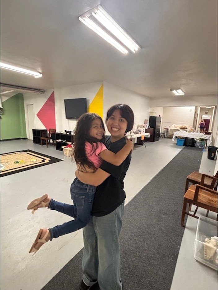 A woman is holding a young girl in an indoor community space. The girl has her arms around the woman and is smiling. The woman is smiling broadly, wearing a black shirt and jeans, while the girl is dressed in a pink top, jeans, and beige shoes. The r