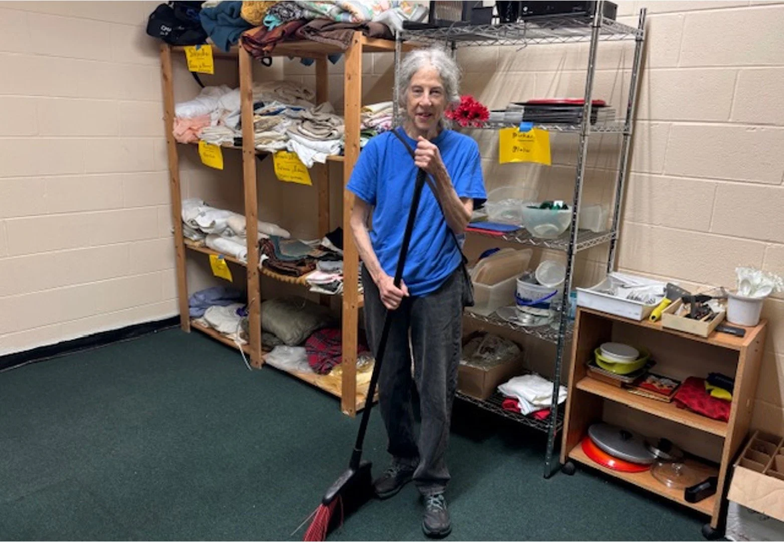 An elderly woman with gray hair is standing in a storage room, holding a broom, wearing a blue t-shirt and dark pants. There are shelves with folded clothes and plastic containers, and the room has beige walls and a green carpet.