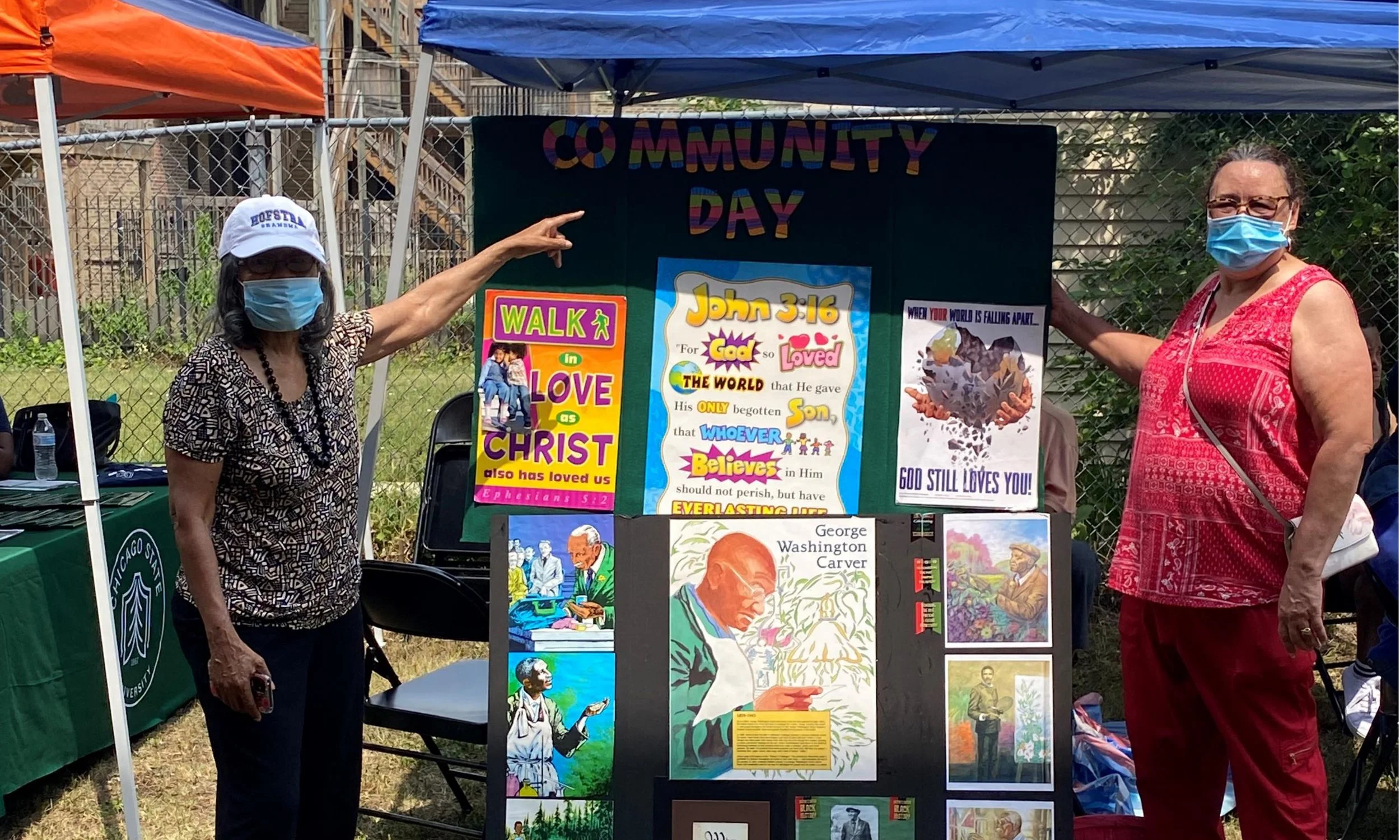 Two women wearing face masks stand in front of a display board at an outdoor community event. One woman points at the board, which features colorful posters and images of John 3:16, George Washington Carver, and other artwork related to faith and com