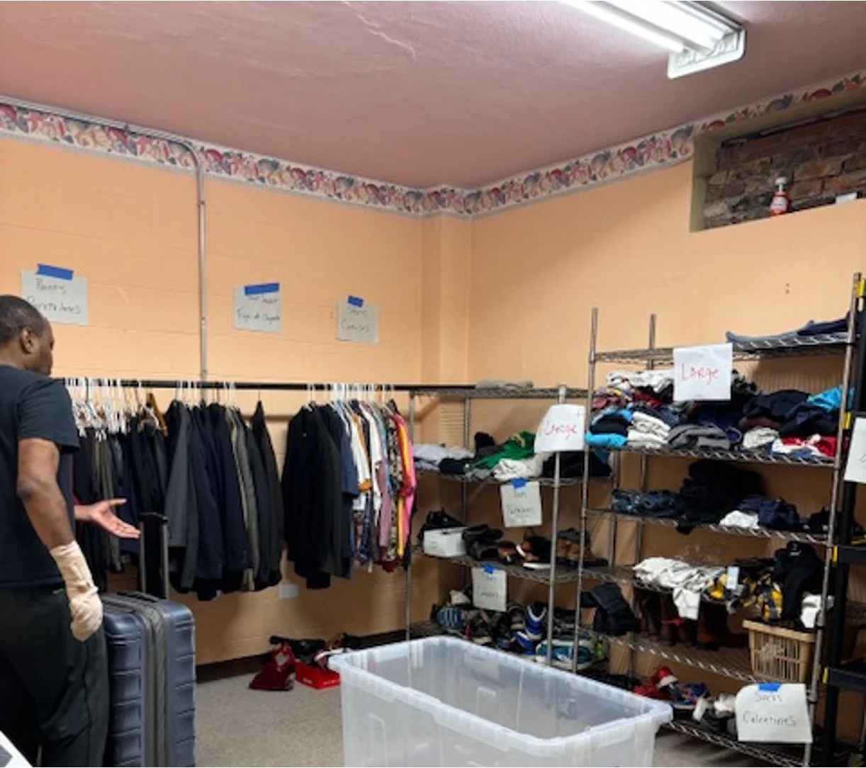 A man with a suitcase and thrift store clothing racks in a room with peach-colored walls and a fluorescent ceiling light.