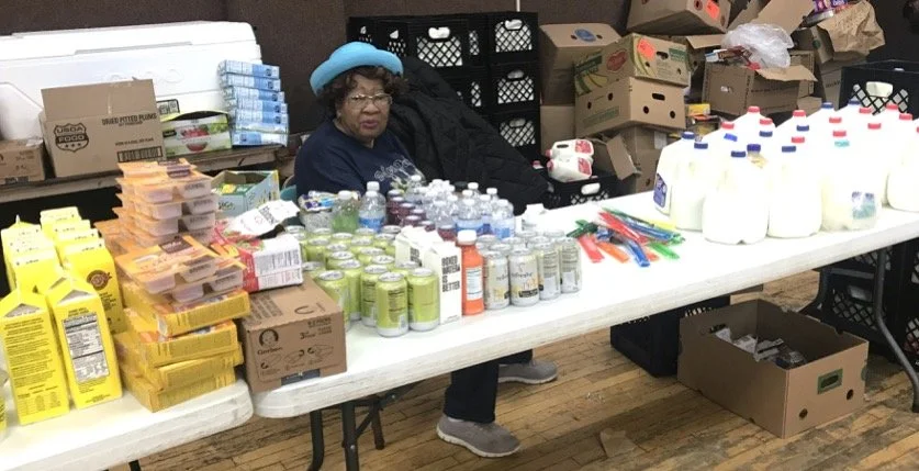 A woman sitting behind a table filled with various food items including canned drinks, bottles of milk, cheese, and snacks, in what appears to be a donation or distribution setup, with boxes and supplies in the background.