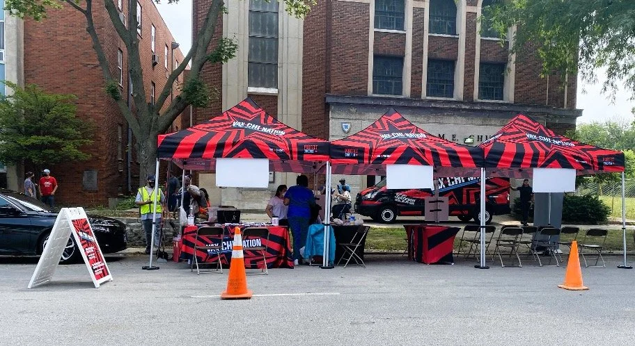 Community event booth set up with red and black tents featuring branding, people gathered around tables, and traffic cones marking the area outside on a city street.