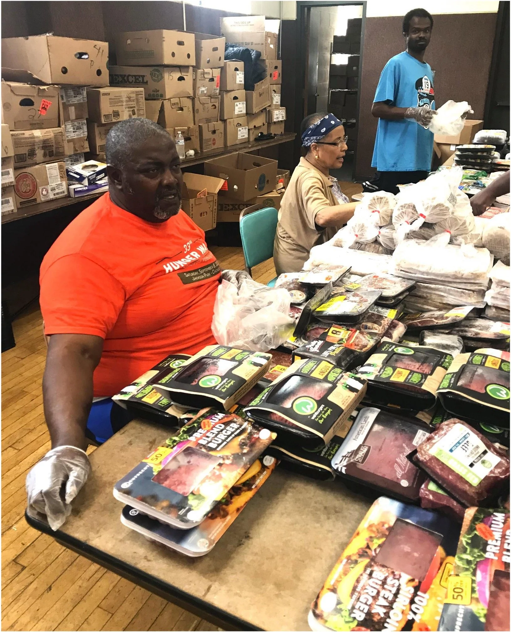 A group of people working at a table packing trays of raw meat at a food distribution event. There are boxes on the shelves behind them and packaging materials on the table.