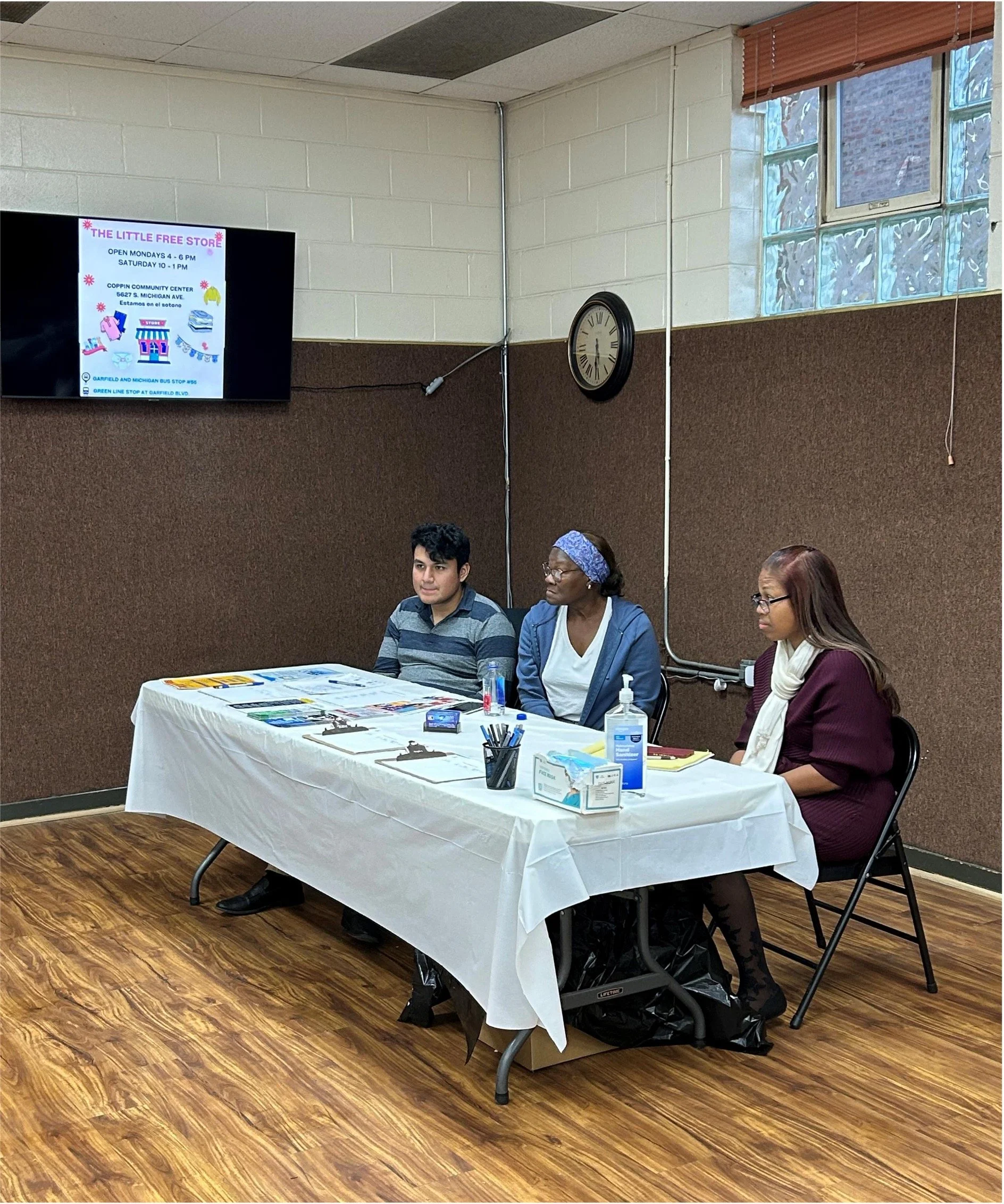 Three people sitting at a table during a presentation or meeting. The table is covered with a white cloth and has various items including pens, notebooks, hand sanitizer, and tissues. Behind them is a brown partitioned wall with a clock and a window.