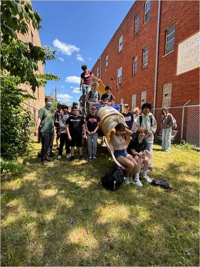 A group of young people gathered outdoors on a grassy area next to a red-brick building, with a slide resembling a cement mixer in the background. Some are sitting and some are standing, and they appear to be enjoying a sunny day.