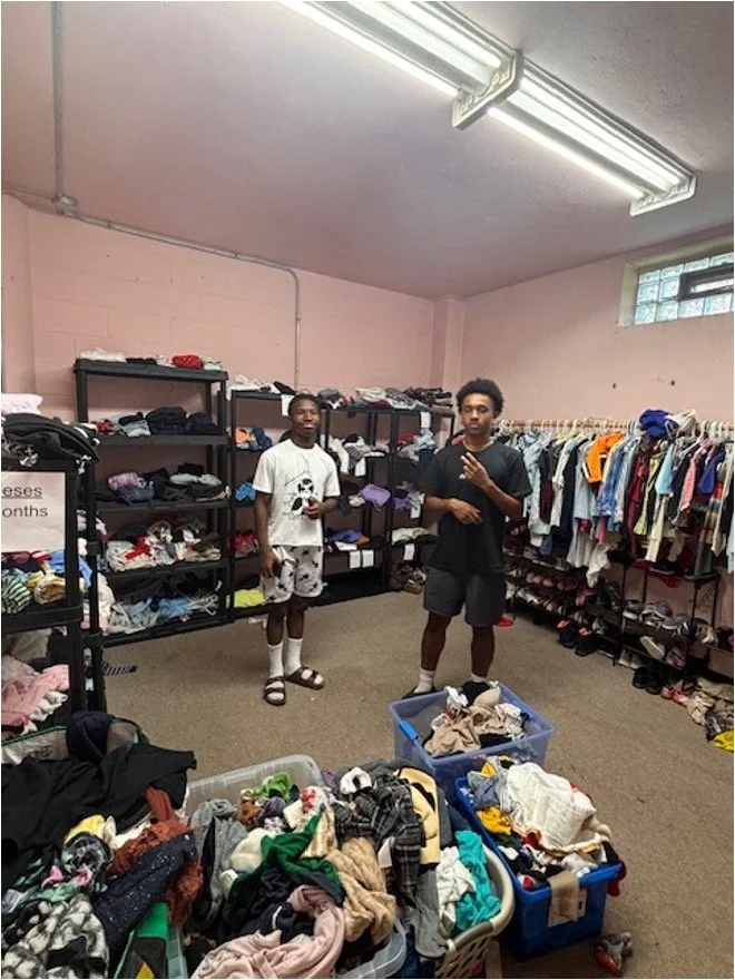 Two young men inside a thrift store or donation center with shelves of folded clothes and hanging garments, as well as plastic bins filled with clothes and fabric in the foreground.
