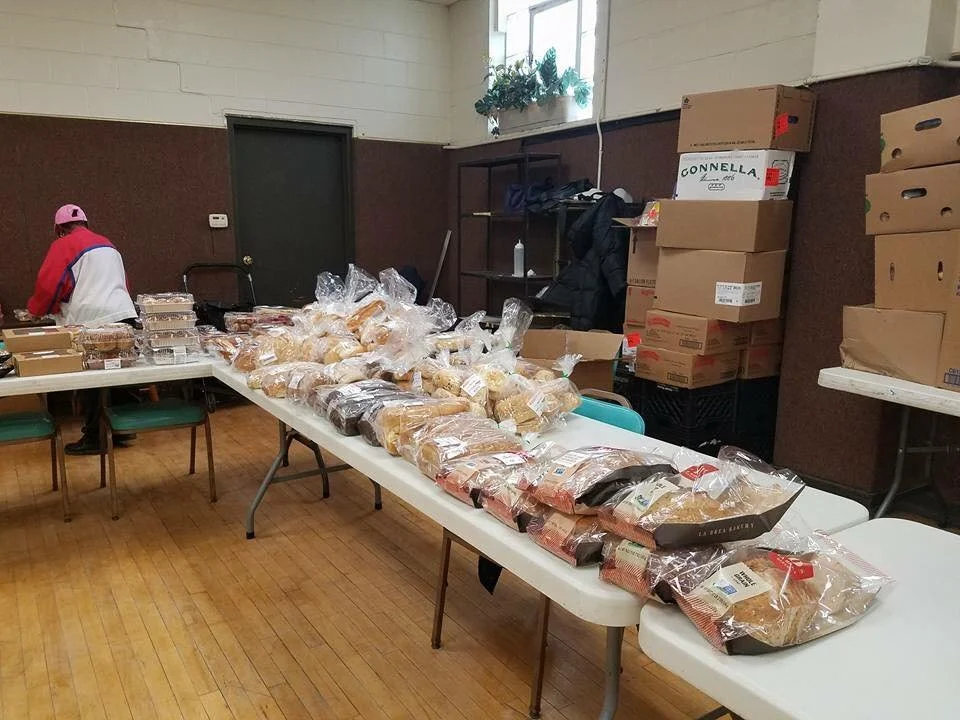 Tables filled with numerous loaves of bread in plastic bags, with boxes and supplies in the background in an indoor setting.