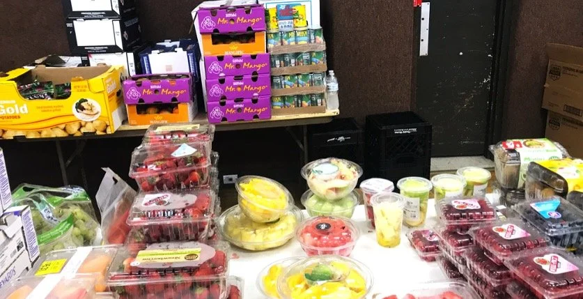 Table filled with containers of cut fruit, including strawberries, grapes, melons, and pineapple, displayed for sale.