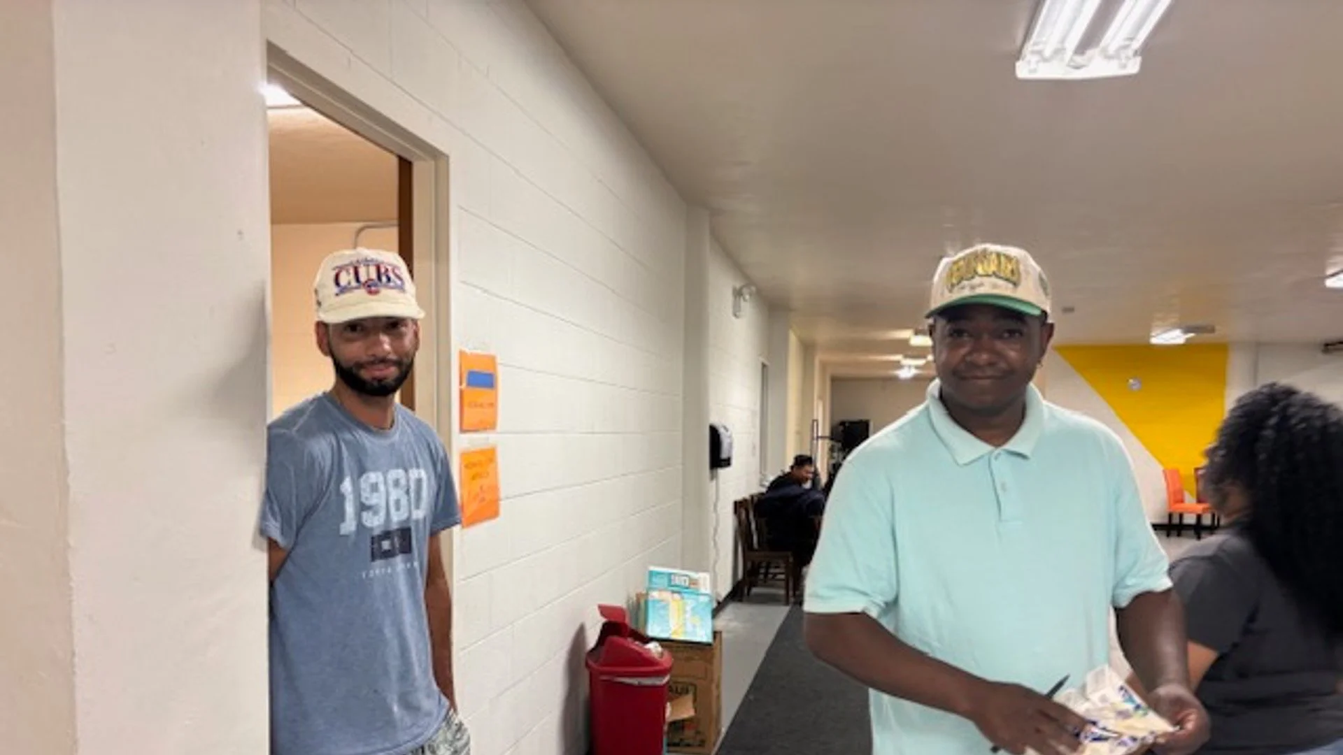 Two men stand in a hallway of a building, one on the left peeking from behind a wall and wearing a Chicago Cubs cap and a t-shirt, the other on the right holding a paper or brochure, wearing a baseball cap and a light-colored polo shirt, with a woman