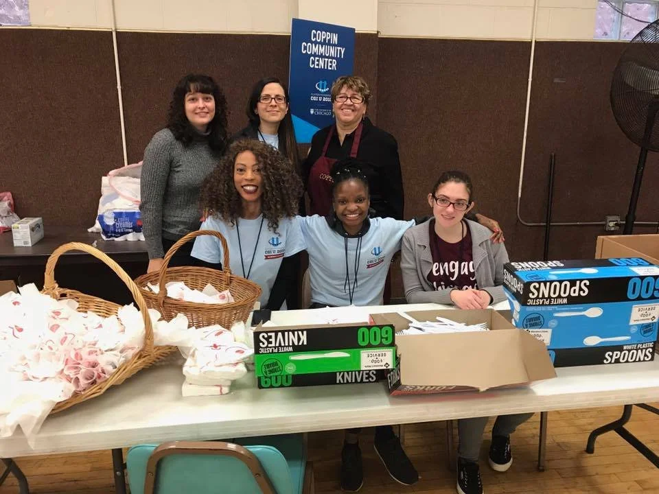 Group of six people smiling behind a table filled with supplies at the Coppin Community Center, with several baskets and boxes of plastic utensils, knives, and spoons.