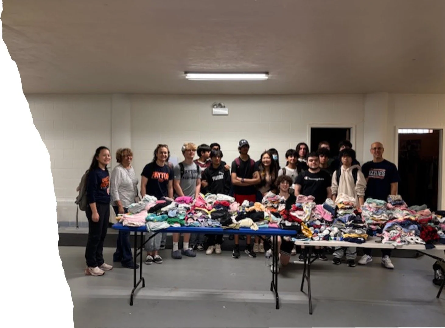 Group of people standing behind tables filled with piles of clothing in an indoor setting.