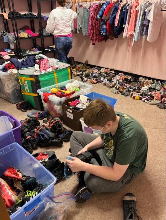 A person sitting on the floor organizing shoes and clothes in a thrift store or clothing donation center, with racks of clothing and shelves of shoes in the background.