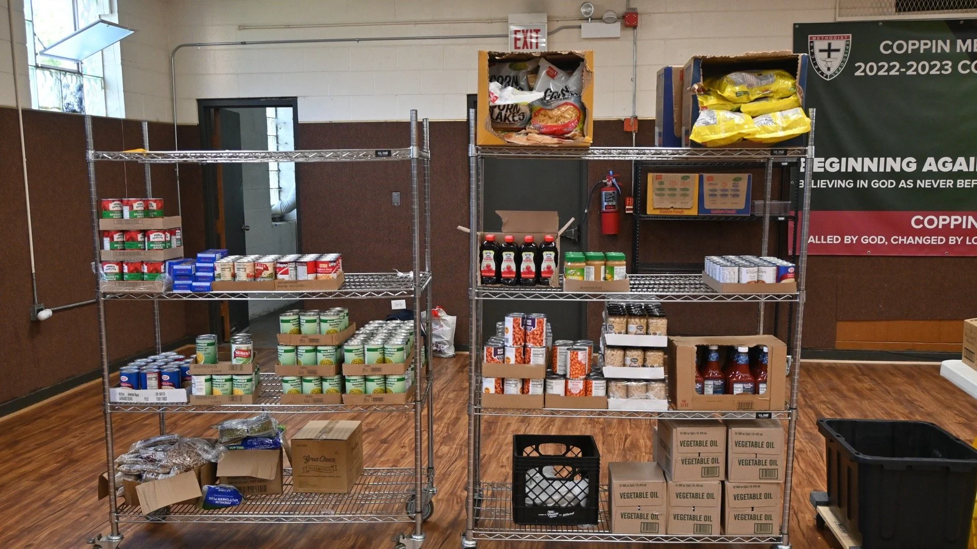 Metal shelving units stocked with canned goods, boxed food items, bottled sauces, and other pantry items in a room with a wood floor and a green banner on the wall.