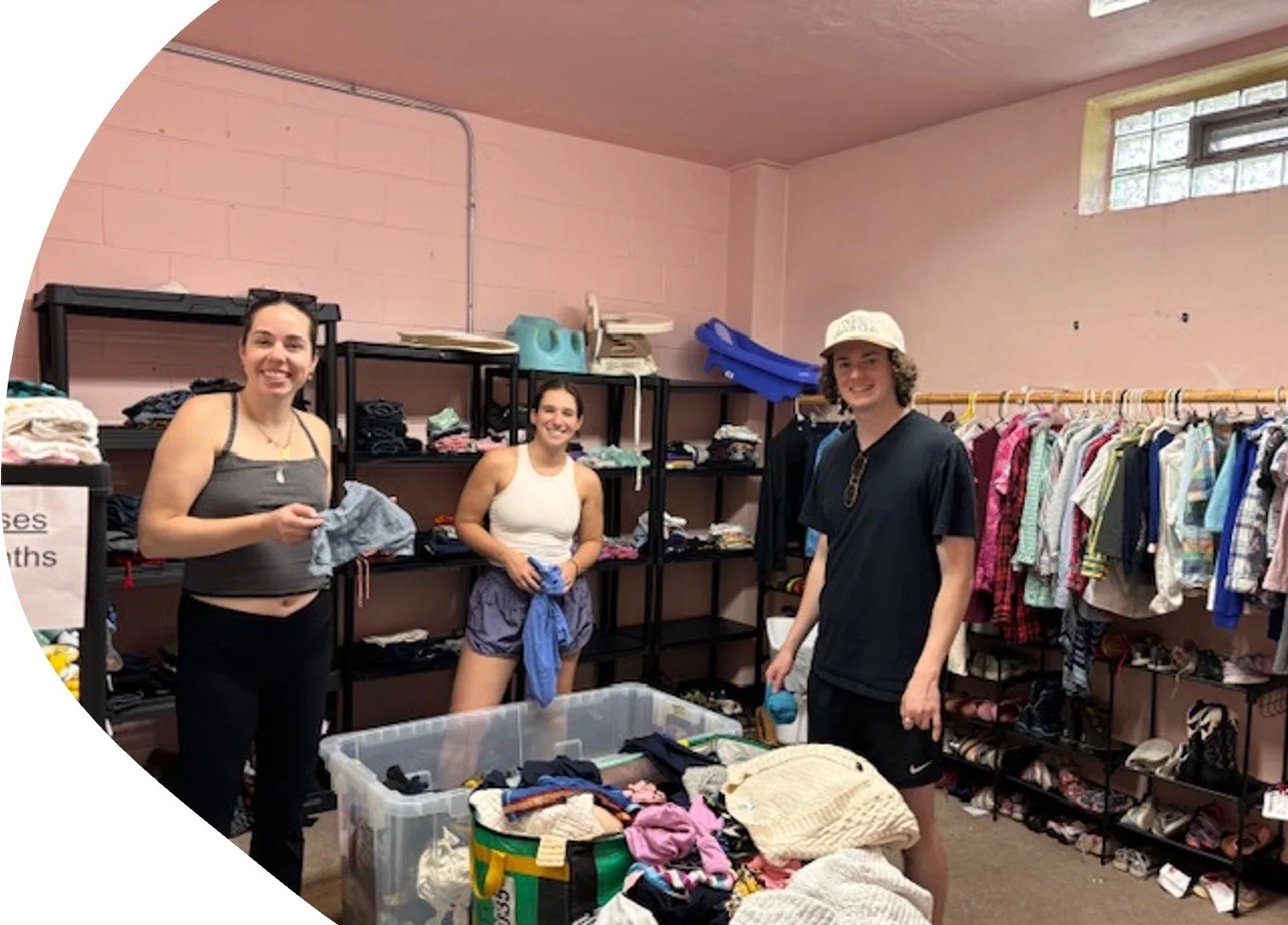 Three young people organizing clothes in a thrift store. Shelves filled with folded clothes and a rack with hanging garments are behind them. A table with piles of clothing and a large plastic bin are in the foreground. The room has pink walls and a 