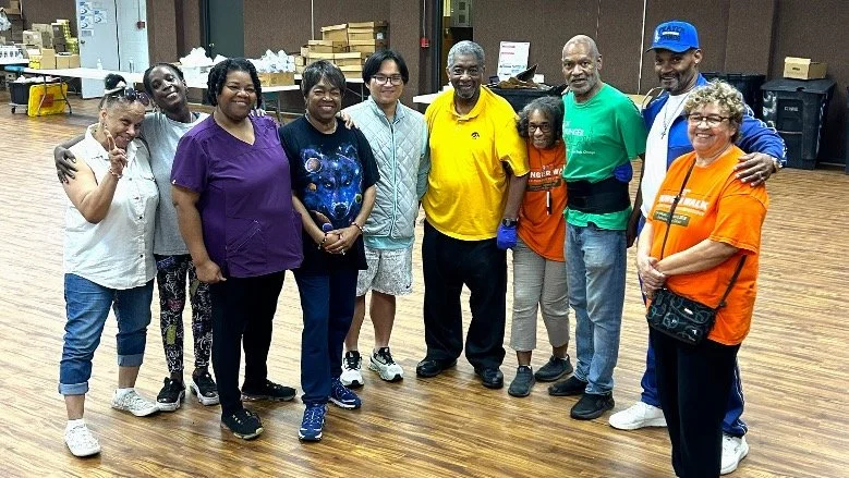Ten people standing together in a room with wooden floors, with some cardboard boxes and equipment in the background. They are smiling and posing for the photo, dressed casually, with a mix of colorful shirts and pants.