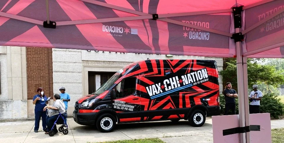 A van with a bold black and red geometric design and the word "VAX-CHATION" on its side parked on a sidewalk under a pink and gray canopy. Several people, including a woman with a scooter and others wearing masks, are standing nearby.