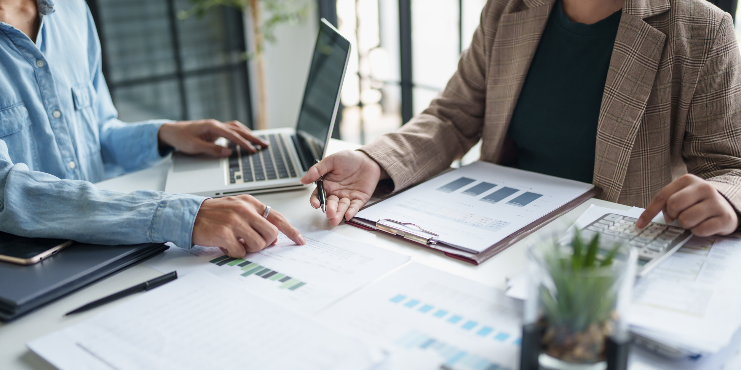 Two people collaborating at a desk with documents, a laptop, and a calculator, discussing charts and data.
