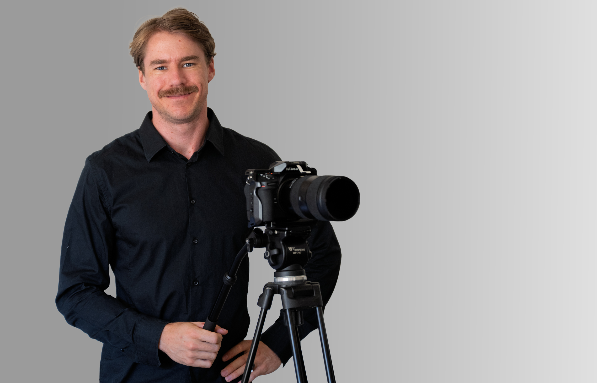 A man with a mustache and light brown hair, wearing a black shirt, is standing next to a professional camera mounted on a tripod against a gradient gray background.