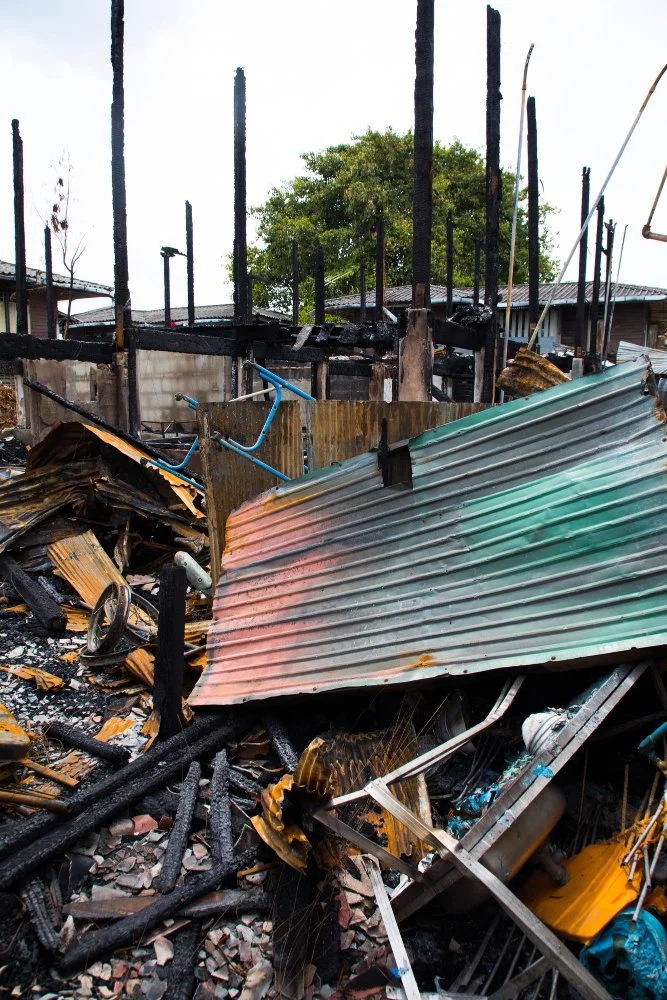 Scene of a burned-down building with charred debris, a partially collapsed corrugated metal roof, and burnt remains of bicycles, with a background of trees and neighboring houses.