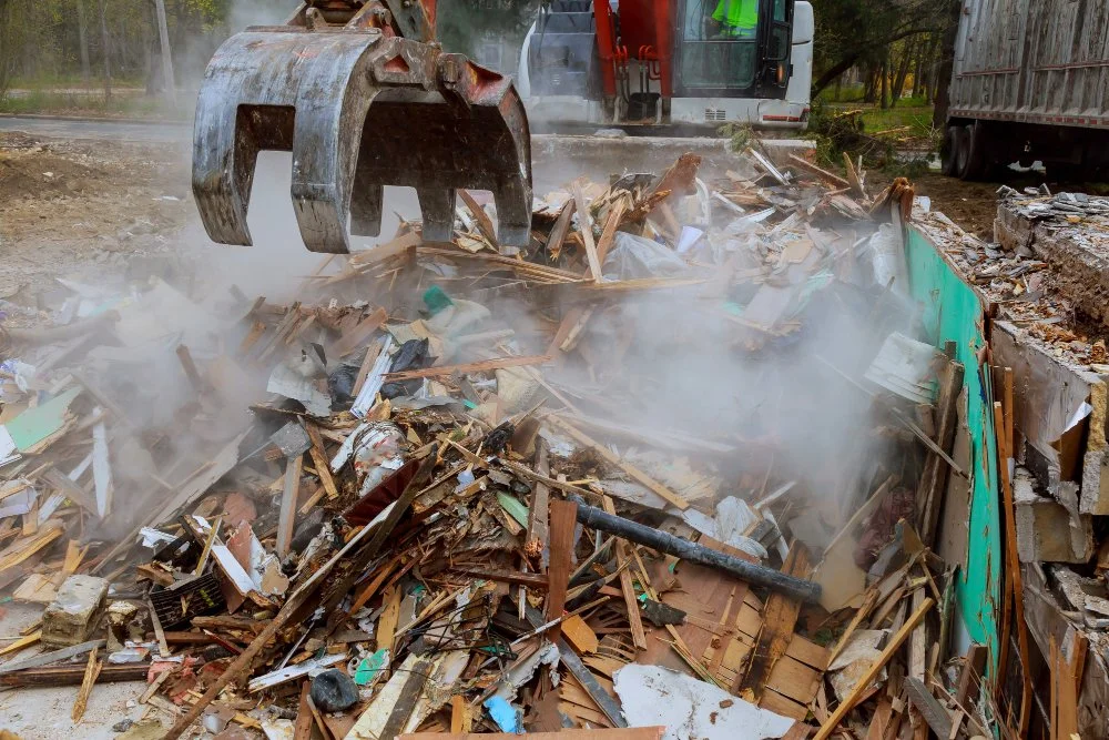 Construction site with a large excavator removing debris and rubble, with dust rising and construction vehicles visible in the background.