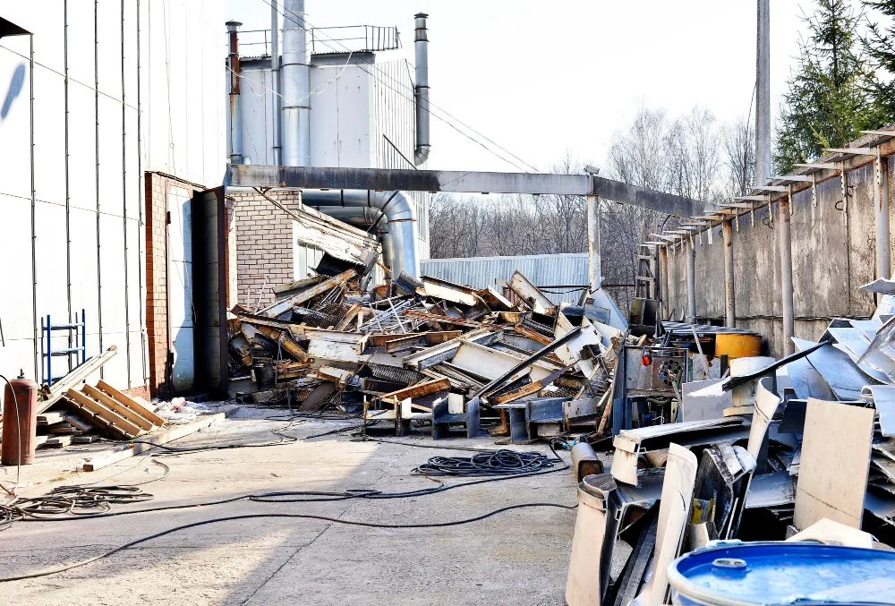 Discarded metal objects and scrap piled up outdoors near industrial buildings.