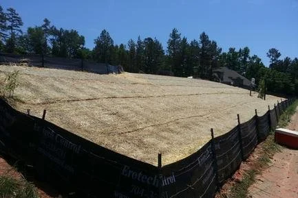 A large plot of land enclosed by black erosion control fabric fencing, with a row of trees and a clear blue sky in the background.