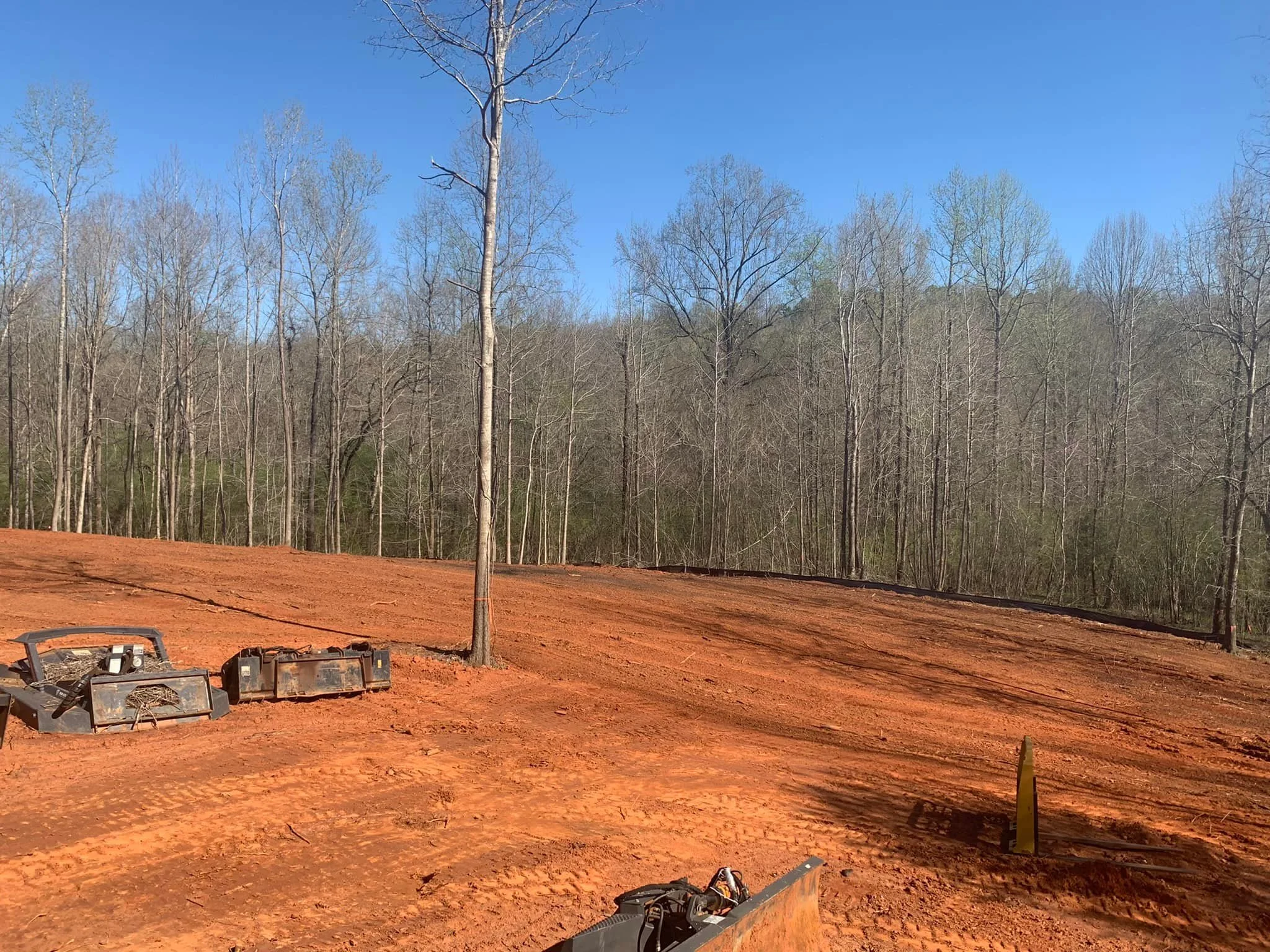 A cleared construction site with red dirt, several tree stumps, and leafless trees in the background under a clear blue sky.