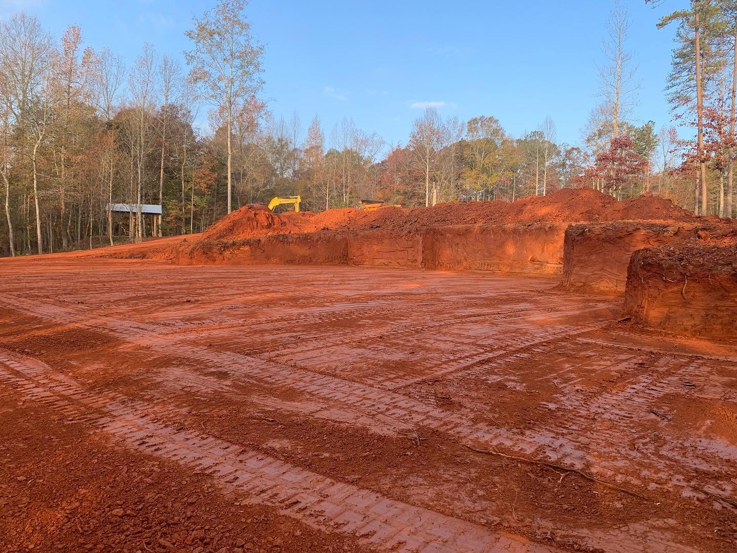 Construction site with a cleared area of red soil and tire tracks, with a yellow excavator and dirt mounds in the background, edged by trees under a blue sky.